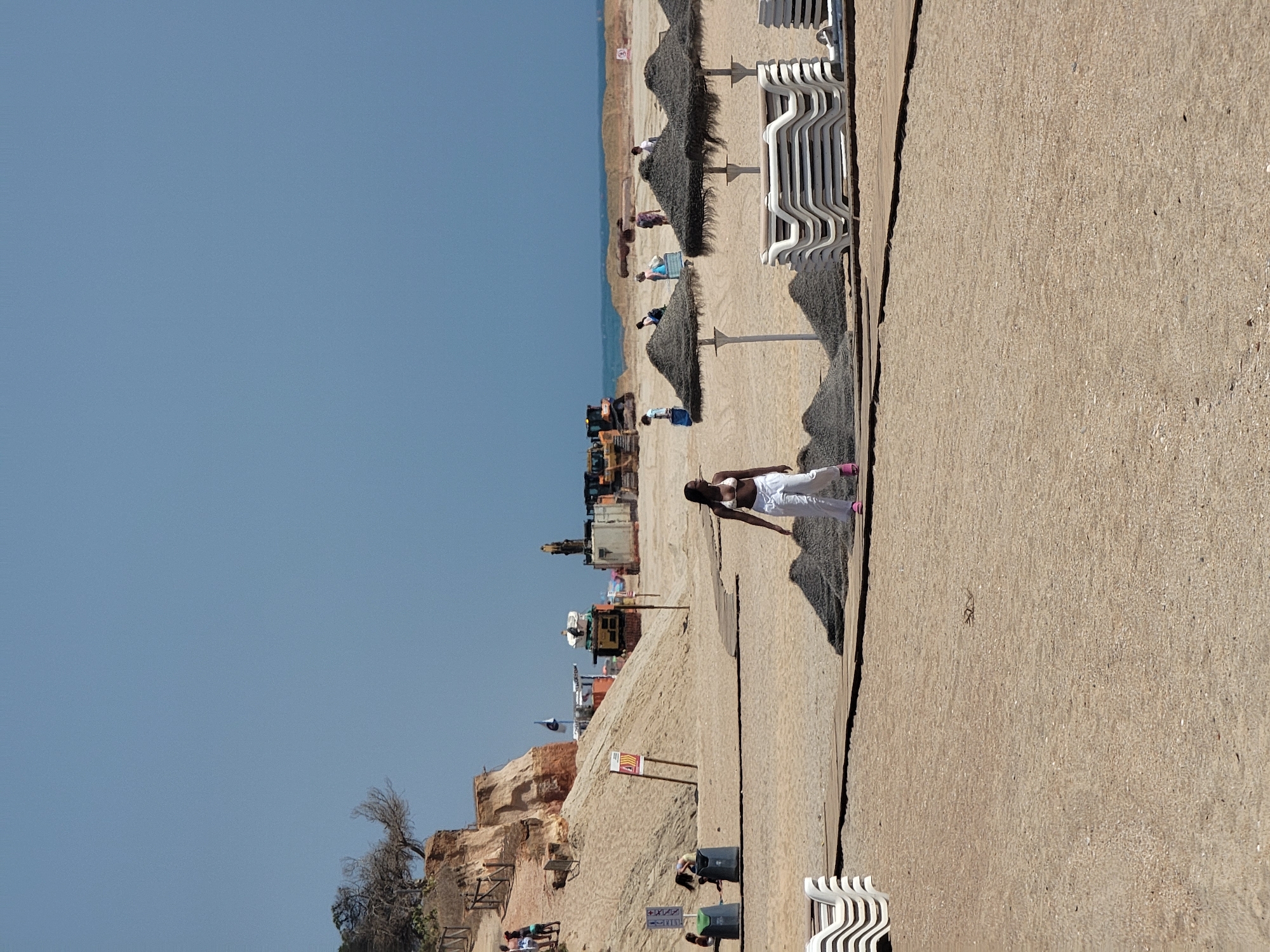The image depicts a construction site on a sandy beach in Quarteira, Portugal. A woman in a white bikini and white pants walks on a wooden path in the foreground. Behind her, stacks of lounge chairs and thatched umbrellas are visible. Further back, construction equipment, including bulldozers, is present. The beach is bordered by a sandy cliff on the left and the ocean in the distance. The sky is a clear blue.