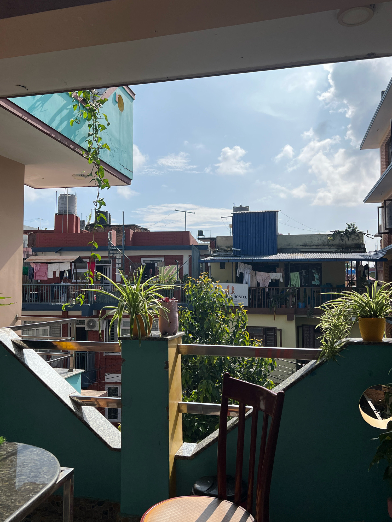 The image depicts a view from a balcony in a city. In the foreground, there is a glass-top table with a metal frame and a wooden chair. The balcony railing is visible, made of metal.
Beyond the balcony, the scene shows a dense urban environment with multiple buildings. The buildings are of varying heights and architectural styles, with some appearing to be residential or guesthouses. Laundry is hung out to dry on some of the balconies and clotheslines. Greenery, including trees and potted plants, is present, indicating a mix of urban and natural elements. The sky is partly cloudy with bright sunlight, suggesting a daytime setting. There are no people visible in the frame. The overall impression is one of a lived-in urban space.