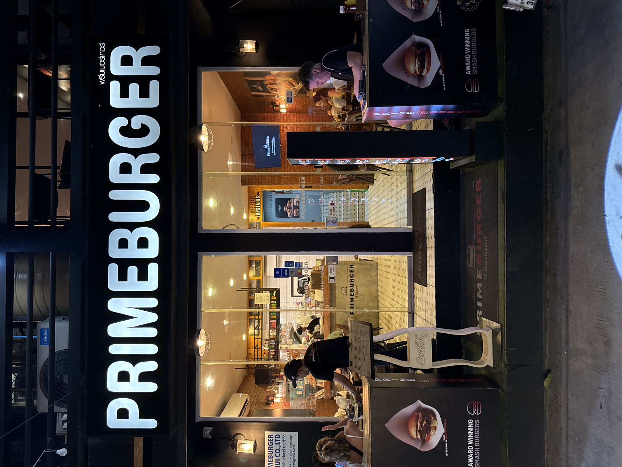 This is a nighttime, outdoor photograph of a PrimeBurger restaurant in Baan Bo Phut, Thailand. The establishment features a prominent vertical sign with "PRIMEBURGER" in large, illuminated white letters. The restaurant's interior is visible through large windows, revealing a brick-lined dining area with warm lighting and patrons seated at tables. Inside, a television screen displays the PrimeBurger logo.
Customers are visible both inside and potentially outside the restaurant. One individual, wearing a black t-shirt and a baseball cap, appears to be an employee, leaning over a counter and interacting with a menu or order screen. Another person, with curly hair, is seated at a table within the restaurant.
Promotional materials are displayed on the exterior of the building. One sign features an image of a smash burger and the text "AWARD WINNING SMASH BURGERS". Another sign indicates "MORE SEATS UPSTAIRS" and "OPEN 11AM - 12AM". Payment methods like Visa are also advertised. A Coca-Cola branded chair is visible outside the establishment. The overall scene suggests a casual dining atmosphere during evening hours. The presence of an air conditioning unit labeled "Midea" is visible on the exterior.