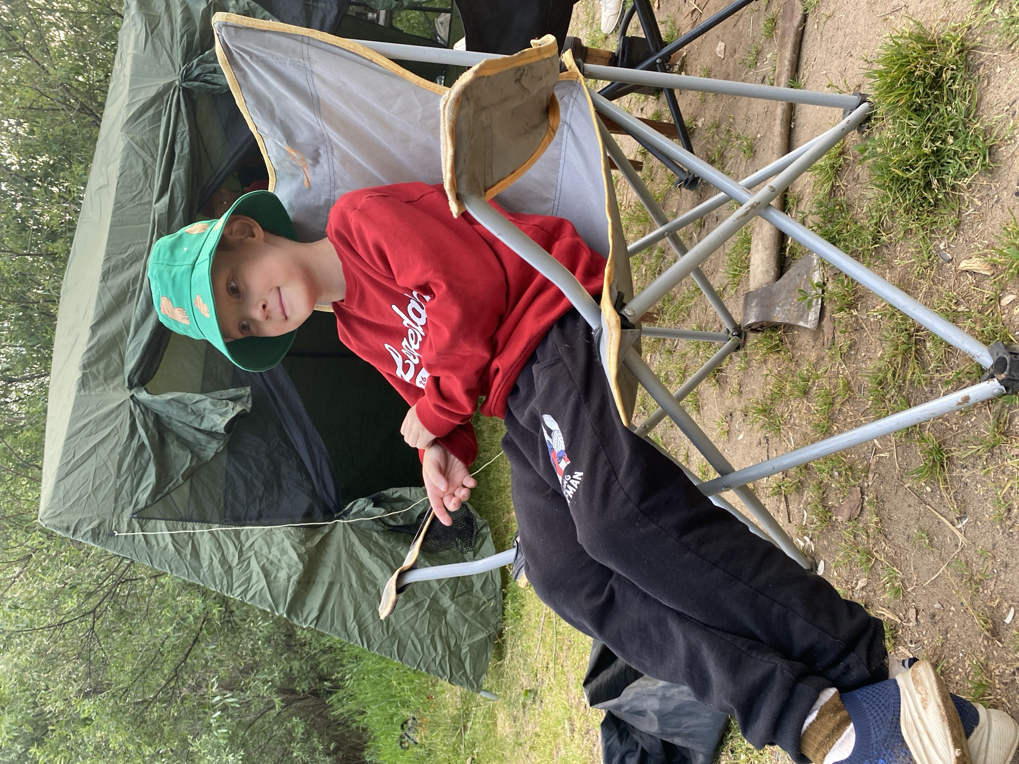 In the city of Olkhovka, Russia, a young boy sits in a folding chair in front of a green tent. He wears a red long-sleeved shirt, black sweatpants with a "Spiderman" graphic, a green bucket hat with orange cartoonish characters, and blue athletic shoes with white soles. His left arm rests on his lap, and his right hand is placed on the armrest of the chair. The tent is partially open, revealing a dark interior. The ground around the campsite is covered in dirt and patches of dry grass. An axe with a wooden handle lies on the ground near the chair. Trees and foliage are visible in the background, suggesting a natural or park-like setting.