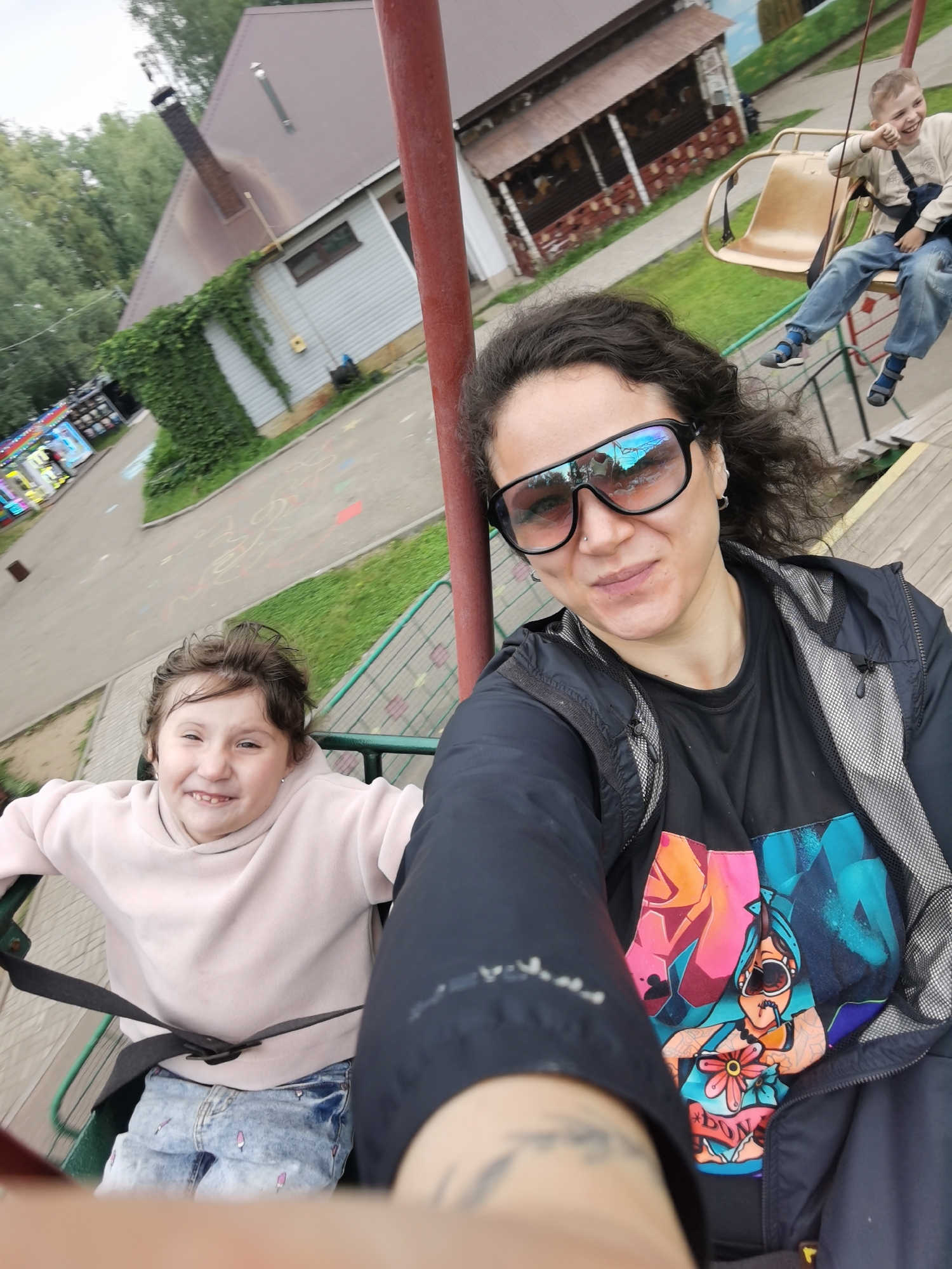 In Chaikovsky, Russia, a selfie is taken on a chairlift ride with two children. In the foreground, a woman with dark, curly hair wears large black sunglasses and a black jacket over a black t-shirt featuring a vibrant graphic of a woman with a cigarette and a flower. To her left and slightly lower, a young girl with brown hair smiles, wearing a light pink hooded sweatshirt and distressed denim jeans adorned with popsicle patterns. Behind them, a young boy in a beige long-sleeved shirt and blue jeans laughs with his arm raised, also seated on a chairlift. The chairlift is a common amusement park ride, with metal framing and beige seats. The background reveals a rustic building with a brown roof and wooden siding, surrounded by greenery and trees, suggesting an outdoor recreational area or amusement park in Chaikovsky.