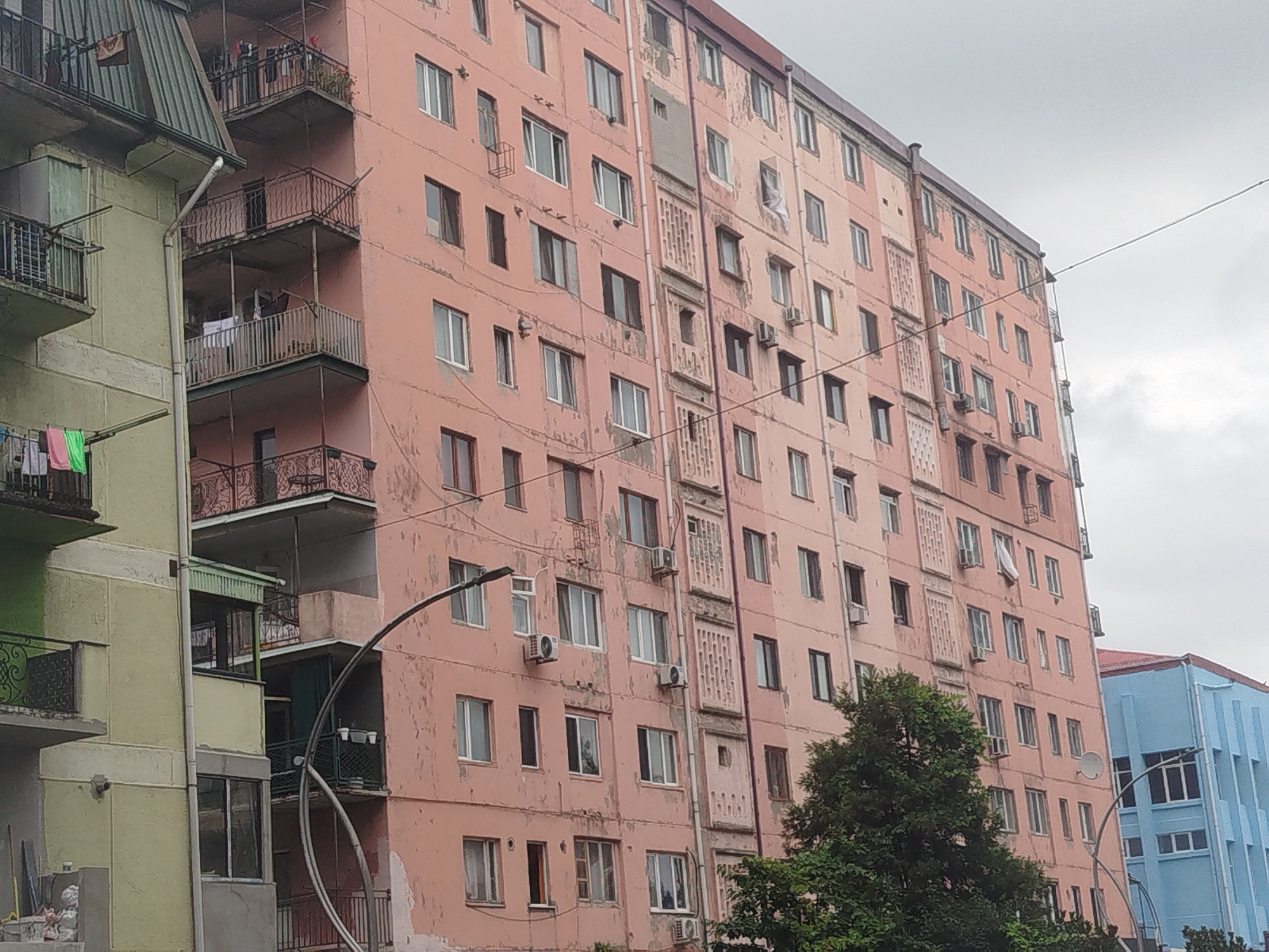 The image shows a view of apartment buildings in Batumi, Georgia, with a pink building dominating the foreground. The pink building is tall and appears to be multi-story, with numerous windows and balconies. Some of the balconies have railings and are adorned with plants or laundry. There are also air conditioning units mounted on the exterior walls of the pink building. Adjacent to the pink building, to the left, is a lighter colored building, possibly green or pale yellow, with similar architectural features. Further to the right, a blue building is partially visible. Trees with lush green foliage are scattered around the base of the buildings. The sky is overcast with clouds. Several wires are visible, suggesting the presence of electrical or communication lines. The overall scene depicts an urban residential area in Batumi.