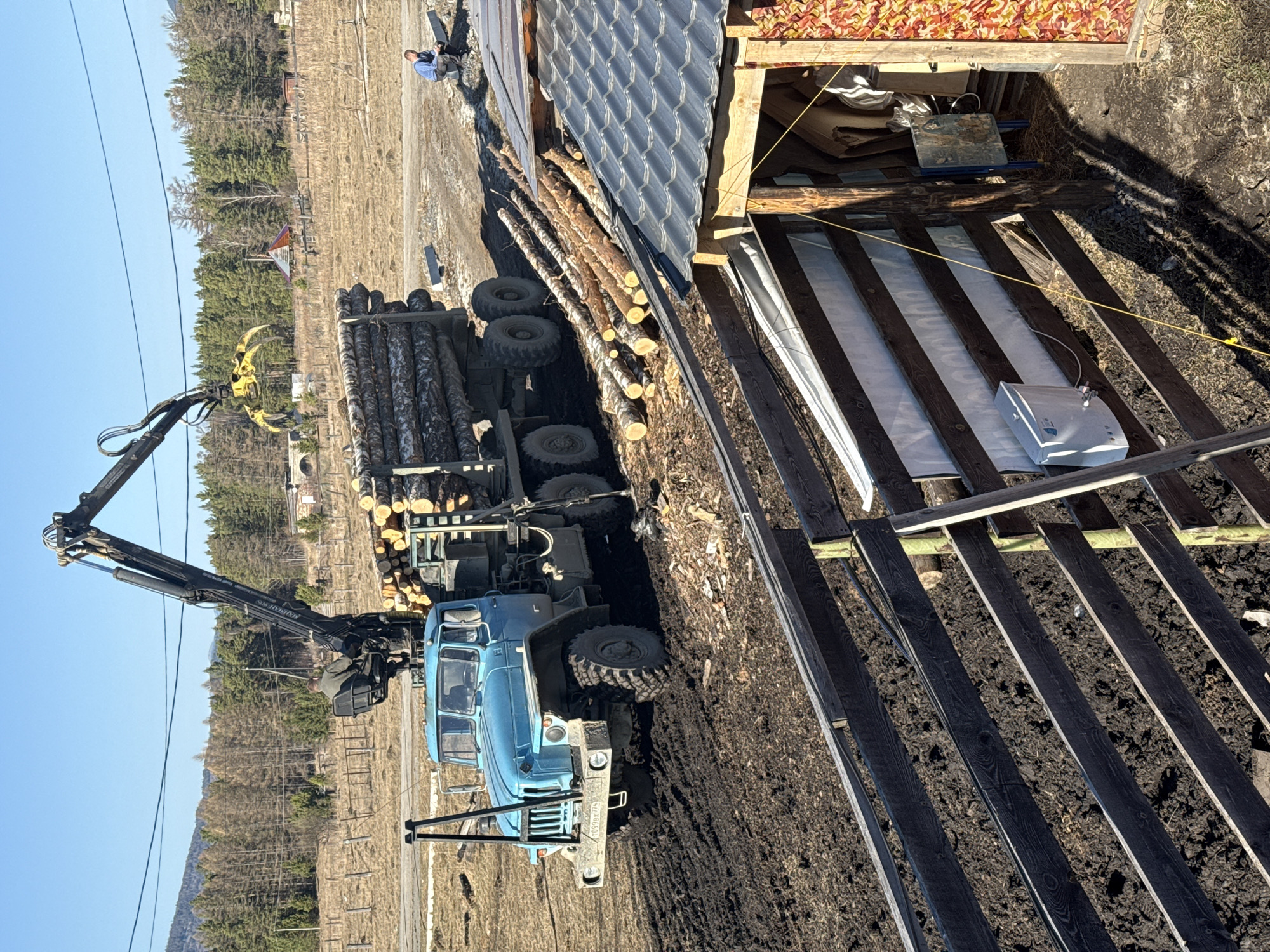 The scene depicts a blue Ural truck loaded with logs, situated on a dirt surface near a partially constructed or renovated wooden structure. The truck is equipped with a hydraulic crane and gripper mechanism, positioned over the load of logs. In the background, a landscape features a mix of open fields and forested areas, likely in a rural or semi-rural setting. In the foreground, a person is seated on the roof of the wooden structure. The environment suggests a work site with ongoing construction or logging activities. The location is Tyulyuk, Russia.