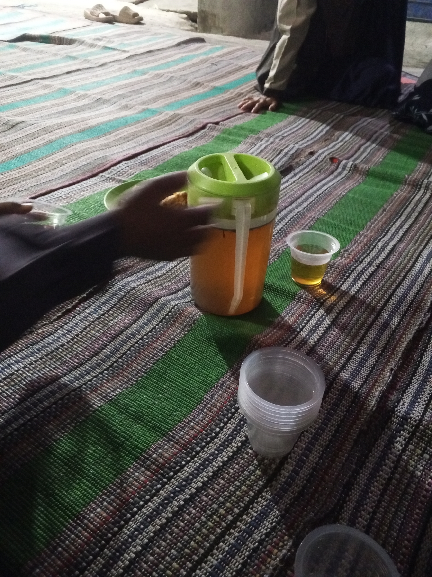 The image depicts an indoor scene, likely a humble dwelling in Ciruas, Indonesia, where a gathering is taking place on a woven mat. The mat features a distinctive pattern of horizontal stripes in shades of green, beige, brown, and black. In the foreground, a stack of clear plastic cups rests on the mat, with another single clear plastic cup containing a yellowish liquid, possibly tea or juice, positioned to the right. A large, orange jug with a green lid and a white spout is prominently placed next to the single cup, suggesting it's the source of the beverage. A hand, partially obscured by shadow, reaches towards the single cup, perhaps to pour more liquid or to pick it up. In the background, a person is seated on the mat, dressed in a dark jacket with beige accents and dark pants. To the far left, a pair of beige slippers are visible. The overall impression is one of a casual, informal gathering, possibly a meal or a break, within a simple living space.