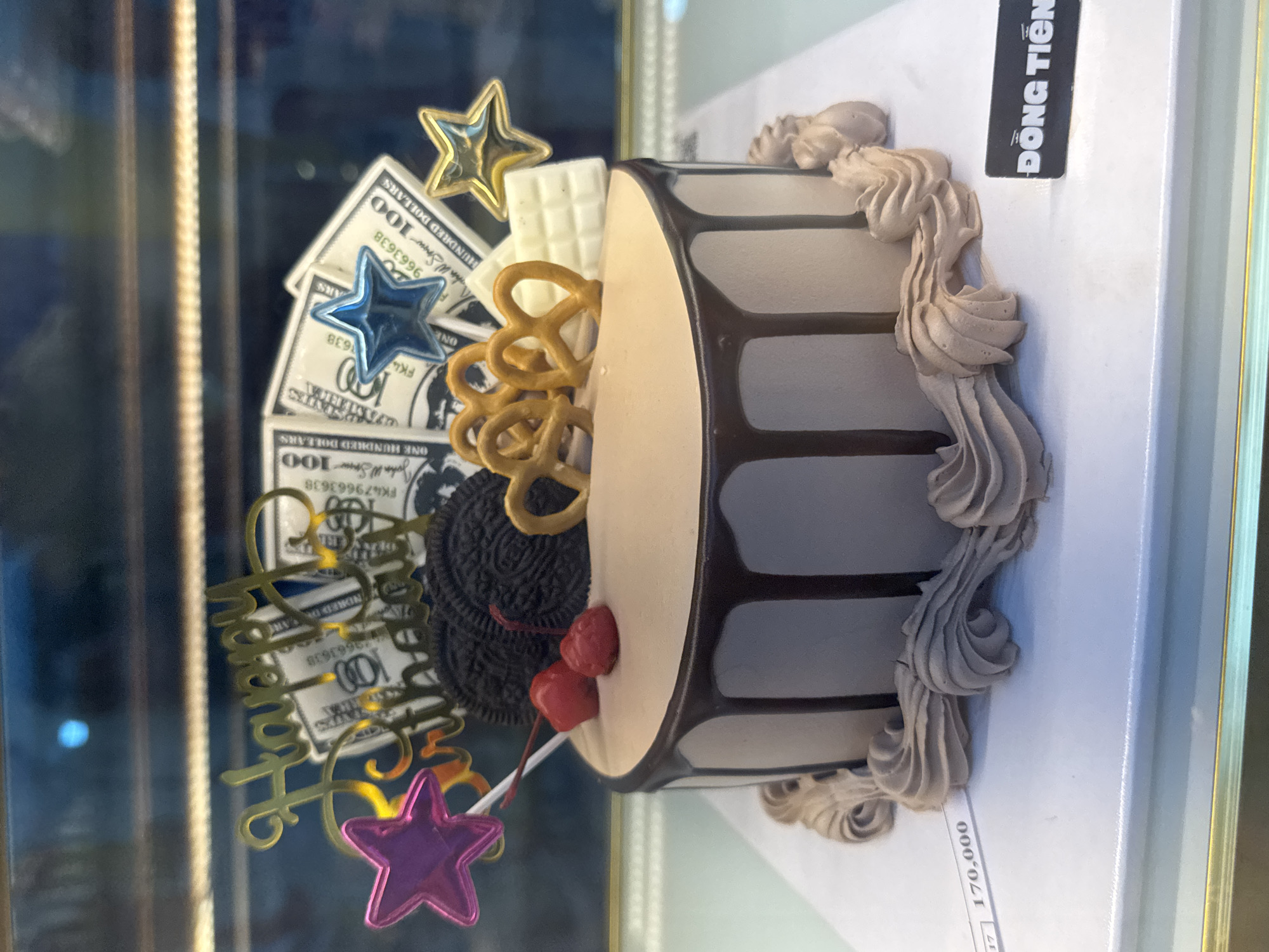 Here is a concise description of the image:
The image shows a decorated birthday cake displayed in a glass case. The cake is round with a beige-colored side, featuring vertical chocolate-colored stripes. It is decorated with frosting swirls around the edges. The top of the cake is adorned with various decorations, including fake hundred-dollar bills, star-shaped ornaments, pretzels, chocolate-covered cookies, and cherries. A gold "Happy Birthday" message is also present.
The cake is likely for sale, as indicated by the "ĐỒNG TIẾN" label and the price "170,000" visible in the lower right corner. Given the language and currency, this is likely in Vietnam. Considering the shop's name DONG TIEN, this image likely comes from Đà Nẵng, Vietnam.