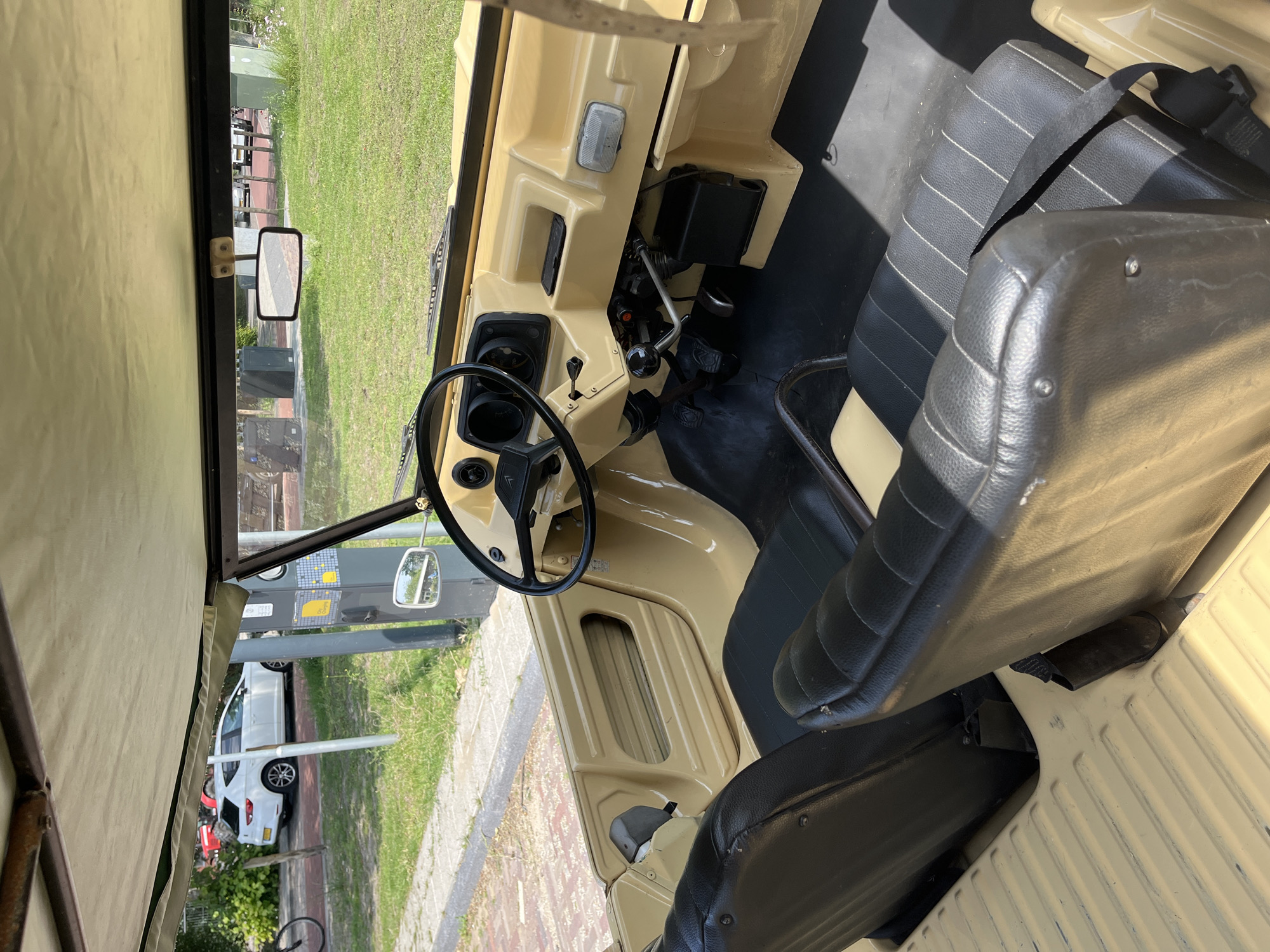The image shows the interior of a beige, open-top vehicle, possibly a small utility car or a dune buggy. The vehicle has two black bucket seats with visible stitching. The dashboard is also beige and features a black steering wheel, a gauge cluster, and various controls. In the background, through the open sides of the vehicle, a paved area with a brick pattern is visible, along with a grassy area and a white car parked in the distance. A street light or utility pole is also present. The overall impression is a casual, outdoor setting.