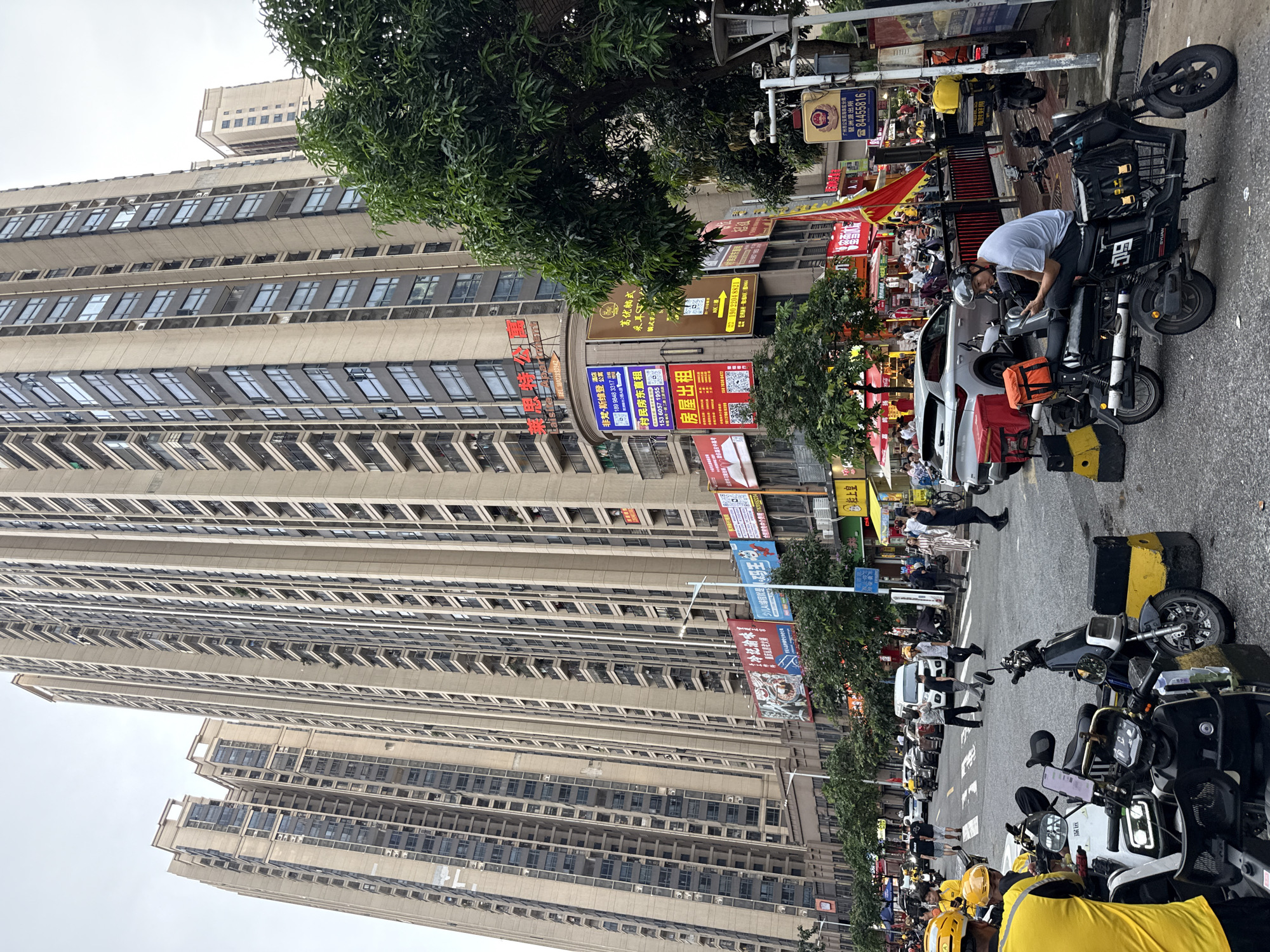 The image captures a street scene in Haizhu District, China, dominated by towering residential apartment buildings. The architecture is modern, with numerous balconies and windows stretching up the facades of the multi-story structures. The street level features shops with illuminated signs and awnings, indicating commercial activity. Several vehicles, including cars and numerous electric scooters, are parked or in motion on the asphalt road. Some of the scooters are equipped with large delivery boxes, suggesting a significant presence of delivery services. People are visible on the sidewalks and crossing the street, some appearing to be pedestrians and others possibly delivery personnel wearing distinctive yellow tops and helmets. A few trees with dense green foliage are interspersed along the sidewalk, providing some natural element to the urban landscape. The sky is overcast, contributing to a diffused lighting condition. A traffic camera and a sign with Chinese characters are mounted on a lamppost on the right side of the frame. The overall impression is one of a bustling urban environment with high-density housing and active commerce.