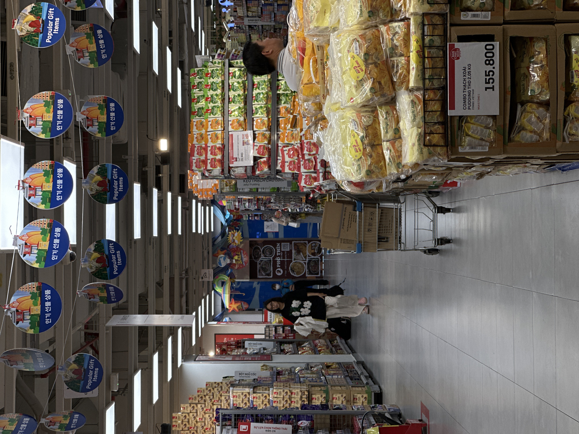 The image shows an interior view of a supermarket aisle in Nha Trang, Vietnam. The aisle is lined with shelves of products, including packaged food items such as cookies and puddings. In the foreground, large stacks of clear plastic bags containing what appear to be puddings are displayed prominently. A price tag on one of these displays indicates "COMBO THẠCH XOÀI PUDDING THD 2.05 KG" with a price of "155.800". A shopping cart is partially visible on the right side of the image. Further down the aisle, a young woman wearing glasses and a black t-shirt with white floral print and beige pants is walking away from the camera. Another person, possibly a store employee wearing a blue uniform, is standing in the background near a food stall labeled "PHỞ THÌN". Overhead, decorative circular ornaments with images of pagodas and boats are hanging from the ceiling, some featuring text in what appears to be Vietnamese or Korean. The supermarket is well-lit with fluorescent lighting.