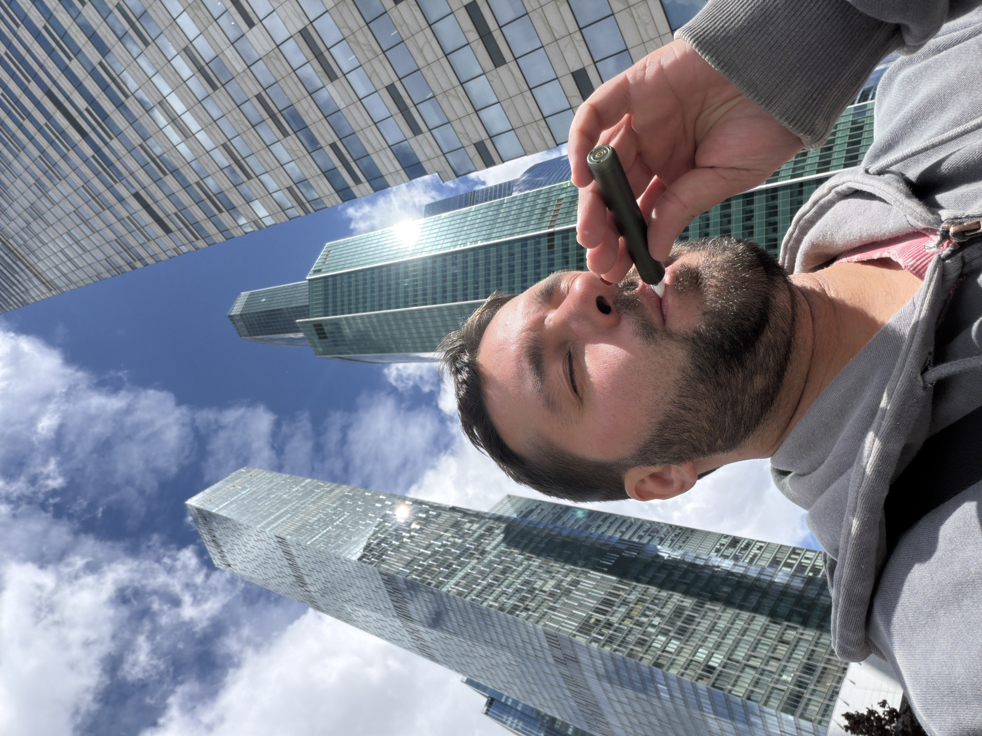 A man is seen from a low angle, appearing to inhale from a small, dark-colored electronic cigarette or vaping device held in his right hand. He has short brown hair and a beard and is wearing a grey hooded sweatshirt. The background consists of multiple modern glass skyscrapers under a partly cloudy blue sky. The buildings appear to be in the Moscow International Business Center, also known as Moscow City.