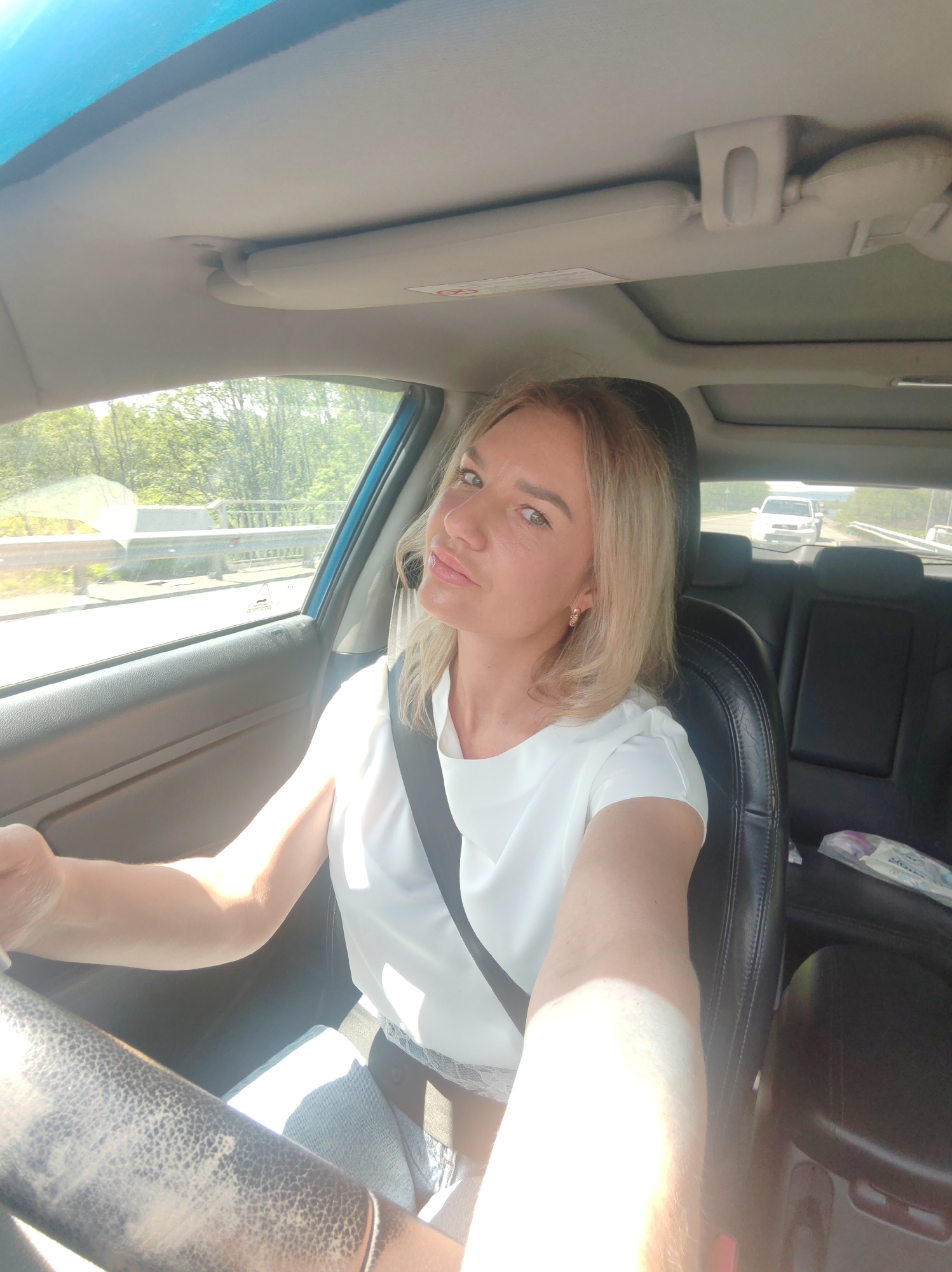 A woman with blonde hair, wearing a white shirt and a seatbelt, is driving a car. She is looking towards the camera and smiling. The car's interior is visible, including the dashboard and seats. Outside the car, trees and a road are visible. The scene appears to be captured in daylight.