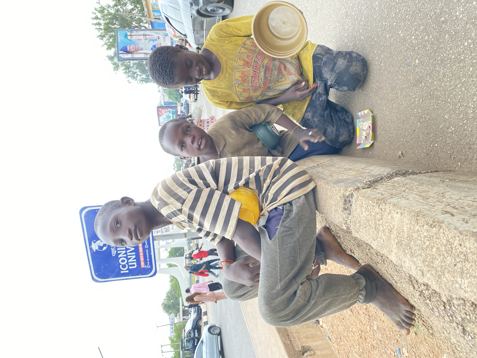 Three barefoot boys sit on a concrete curb on a roadside in Jos, Nigeria, begging for money. One boy wears a striped shirt and sits closest to a blue sign reading "ICONIC UNIV STUDY WITH CLASS RE". Another boy wears a brown shirt, and the third wears a yellow shirt. The yellow-shirted boy holds up a bowl. In the background are cars and people.