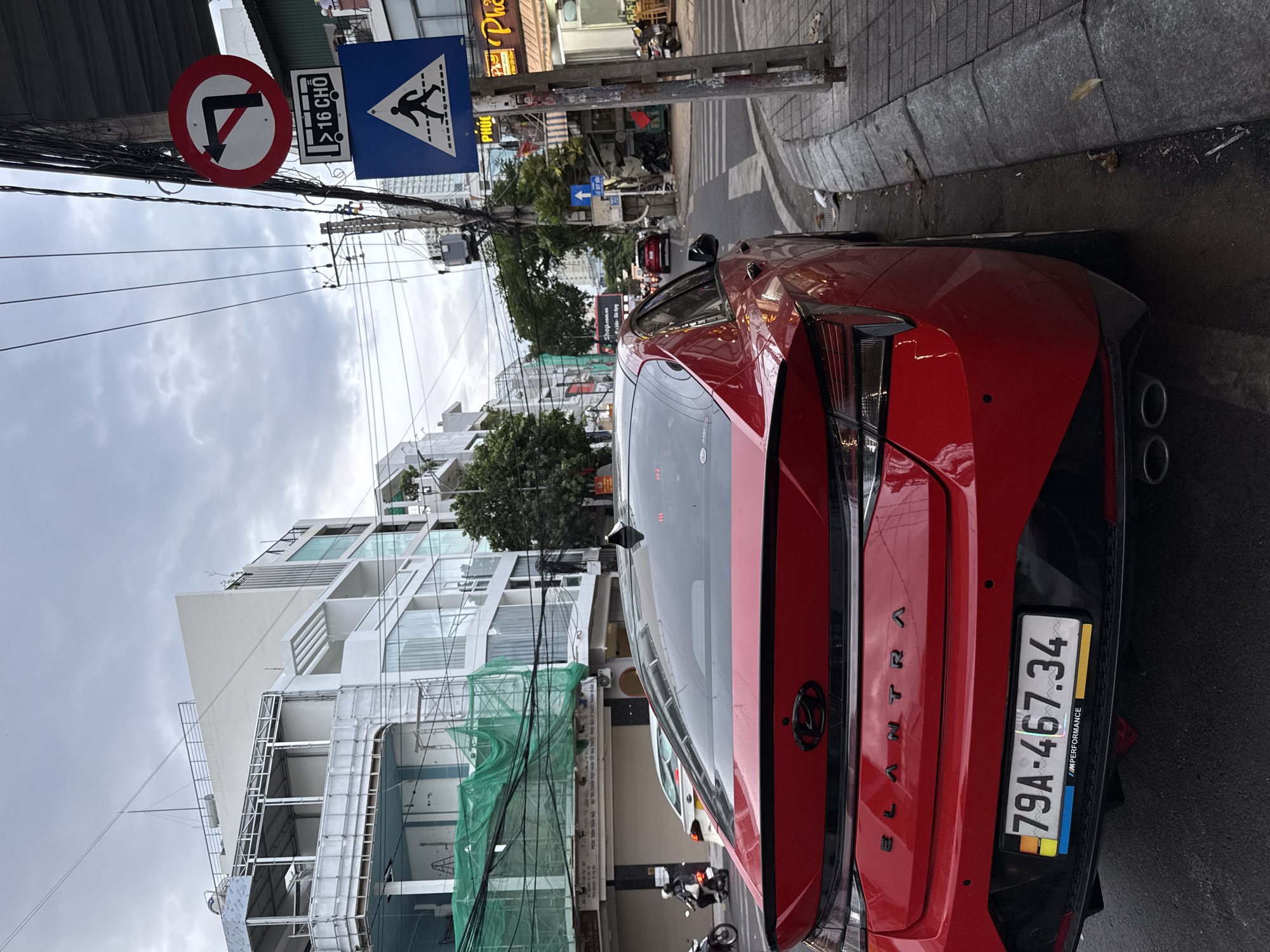 The image shows a red Hyundai Elantra parked on the right side of a street in Nha Trang, Vietnam. The car is facing away from the camera, and its license plate reads "79A-467.34". The street is lined with buildings, some of which appear to be under construction with green netting covering scaffolding. There are traffic signs visible, including a "no left turn" sign and a pedestrian crossing sign. A few motorcycles and other cars are on the street. The sky is overcast. The overall scene depicts a typical urban streetscape in Vietnam.