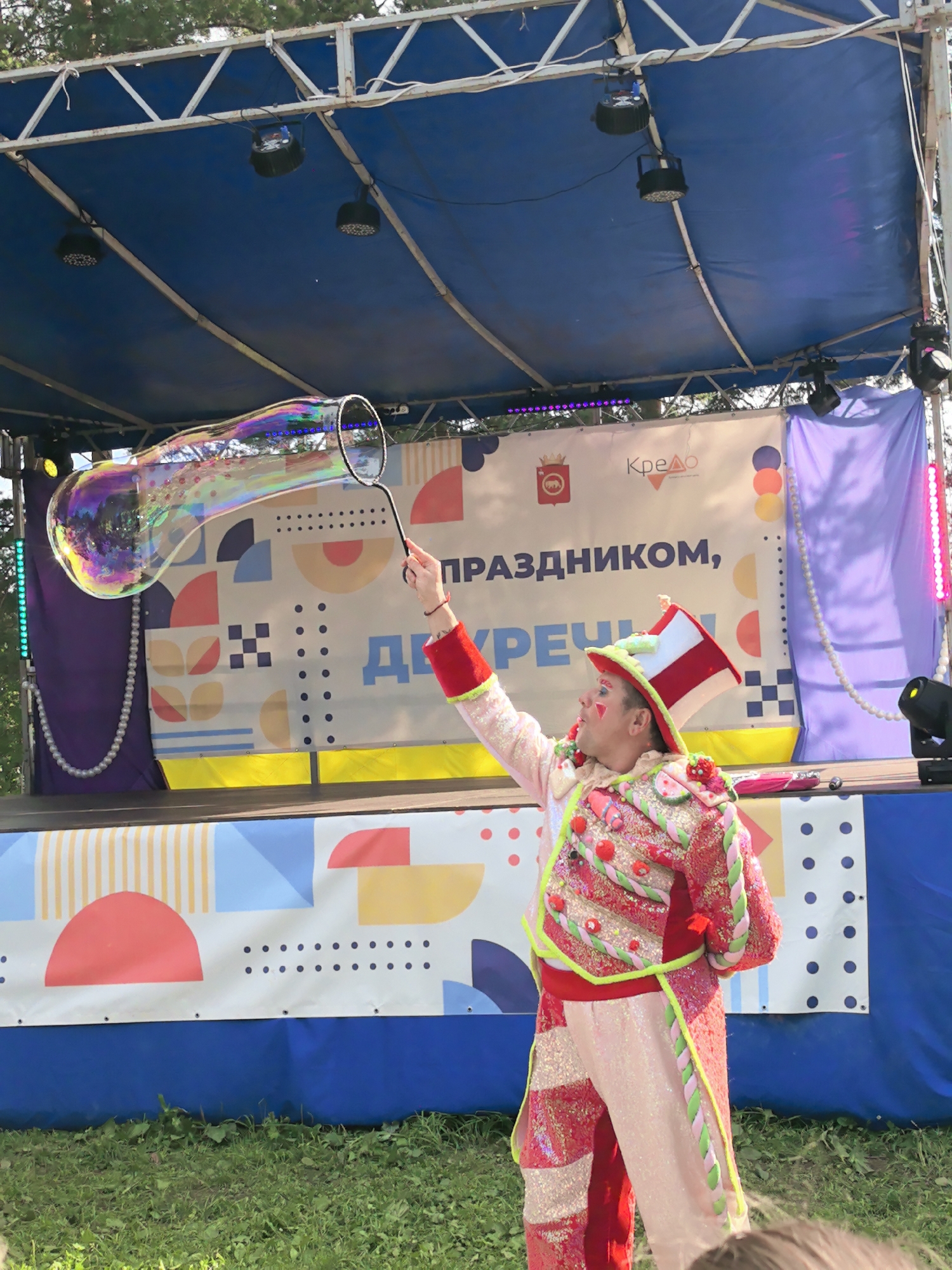 In this image, a performer in a colorful, sequined costume and a tall, striped hat is creating a large, iridescent bubble on an outdoor stage. The performer is holding a wand with a loop, from which the bubble has just emerged. The bubble is elongated and stretched, reflecting the bright lights and colorful backdrop behind the performer.
The stage itself is decorated with a banner featuring geometric shapes and text in Russian. Visible text includes "Кредо" and what appears to be "ПРАЗДНИКОМ," suggesting a celebratory event. There are also what look like the coat of arms of a municipality and colorful circles on the banner. The stage is covered by a blue tarp, and lights are suspended above. The overall setting appears to be outdoors during the daytime, with hints of trees in the background and green grass in the foreground. The performer's bright costume and the playful nature of bubble-making suggest a cheerful and festive atmosphere, likely at a public event or festival in Gorniy, Russia.