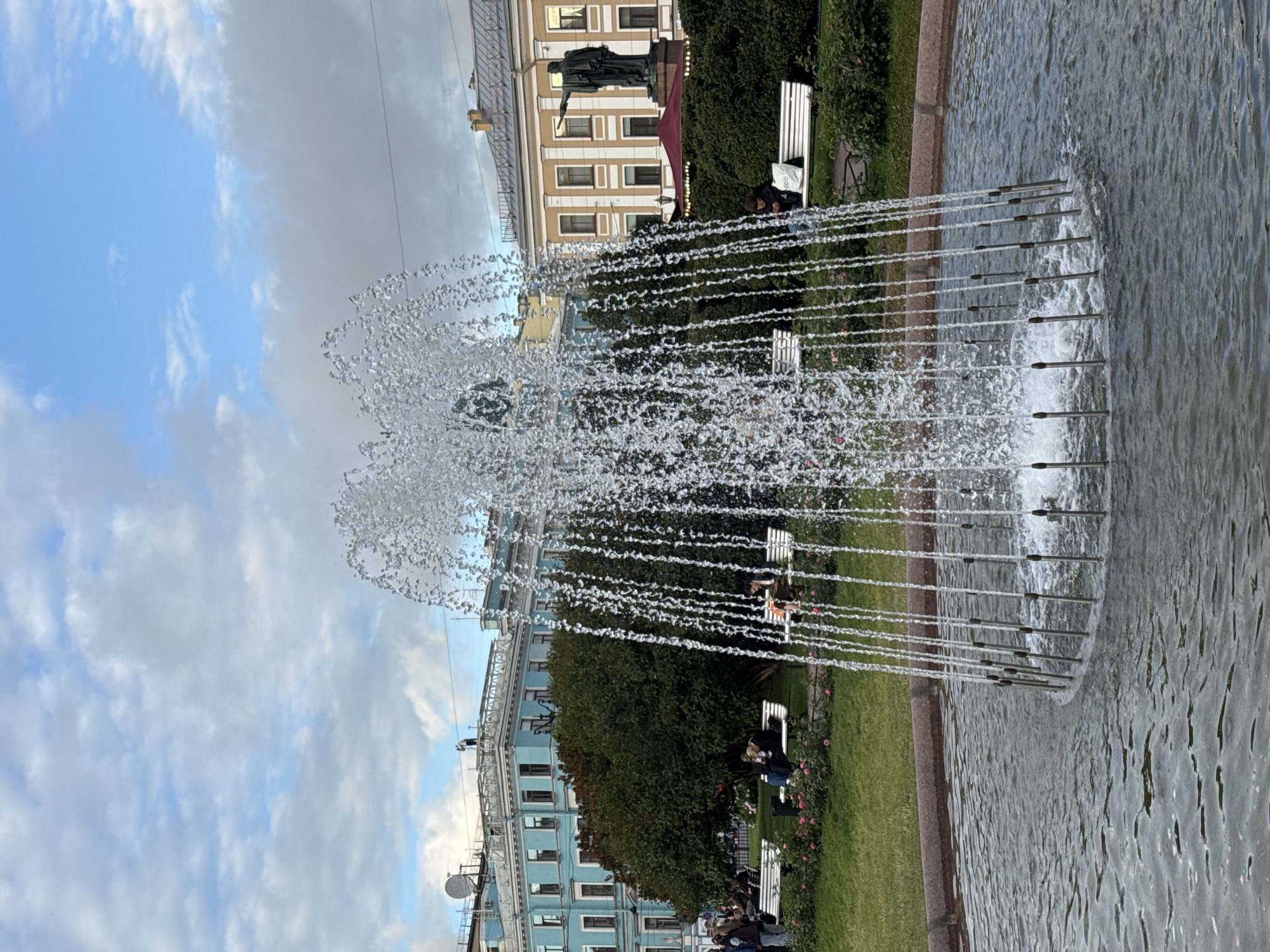 The image shows a fountain in a city square in Saint Petersburg, Russia, likely near Palace Square, given the architectural style of the buildings. The fountain is in the foreground, with water spraying upwards and cascading down in thin streams. The water creates a shimmering effect against the backdrop of buildings and the sky.
In the mid-ground, there are several white benches with people sitting on them. Some individuals are looking at their phones, while others are engaged in conversation or simply enjoying the ambiance. A lush green lawn surrounds the fountain and benches, with some pink flowers adding a touch of color. Behind the lawn, there are several trees, and beyond that, buildings with classical architectural features, including ornate facades and arched windows.
On the right side of the image, a statue of a prominent figure, possibly a historical or literary character, stands tall against the sky. The statue appears to be made of bronze and is positioned on a pedestal. The sky is partly cloudy, with patches of blue visible, indicating a temperate day. The overall scene evokes a sense of tranquility and urban leisure.