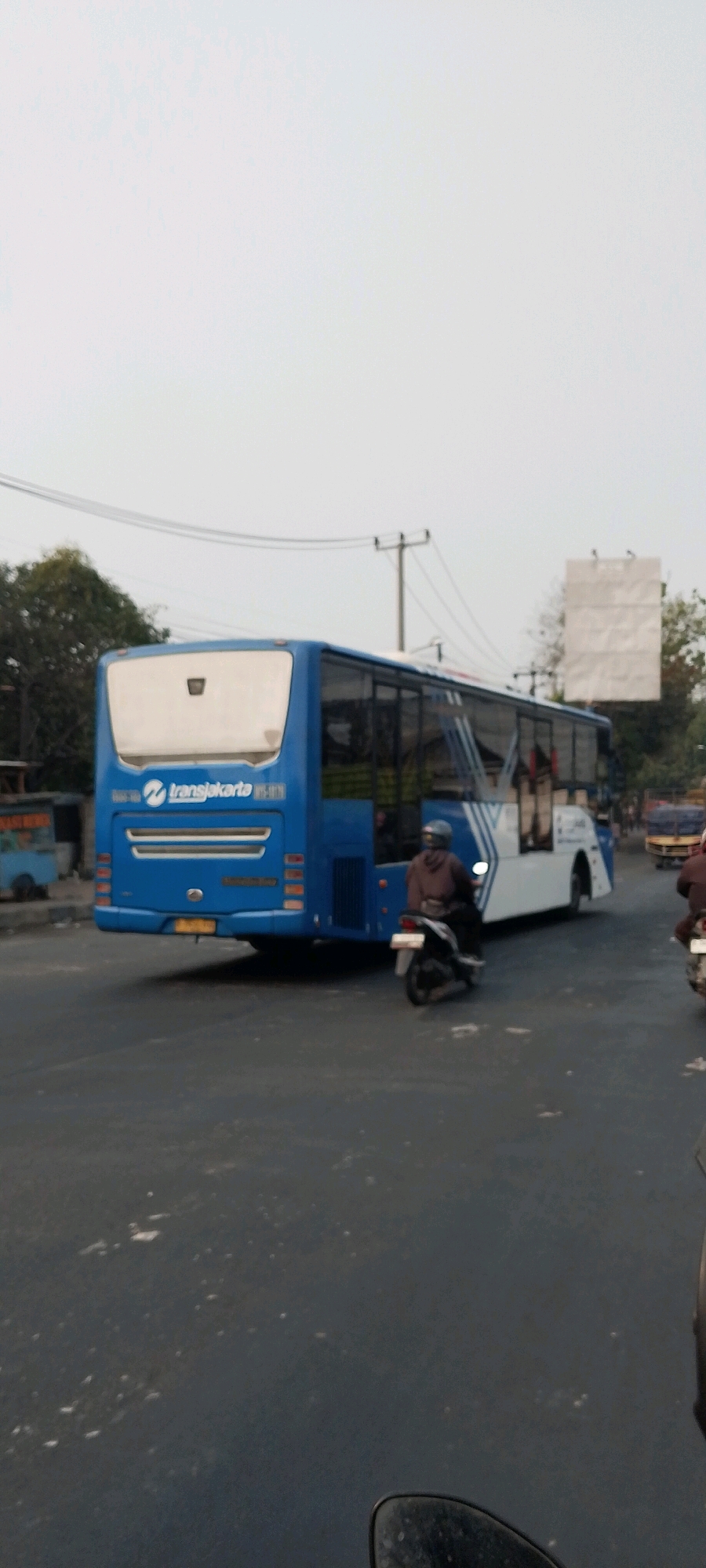 An undated, vertical, medium-shot, eye-level photograph captures a blue TransJakarta bus with a white rear section and a white trailer, moving on a paved road under an overcast sky. The bus, identified by the "transjakarta" logo and text "BS-109" on its rear, is positioned on the right side of the frame, with its front heading towards the left. A motorcycle with a rider wearing a brown jacket and helmet is to the right of the bus, also moving towards the left. The rider's face is obscured by the helmet. The road is dark asphalt with visible white markings. Trees and electrical poles are visible in the background. On the left side of the road, there appears to be a small roadside stall with a blue and white structure. The sky is a uniform light gray, suggesting diffused light conditions. Based on the presence of TransJakarta buses, commonly found in the Jakarta metropolitan area, and the visual cues of urban infrastructure, the scene is likely situated within the city of Bekasi, Indonesia, a major satellite city within the Greater Jakarta region.
