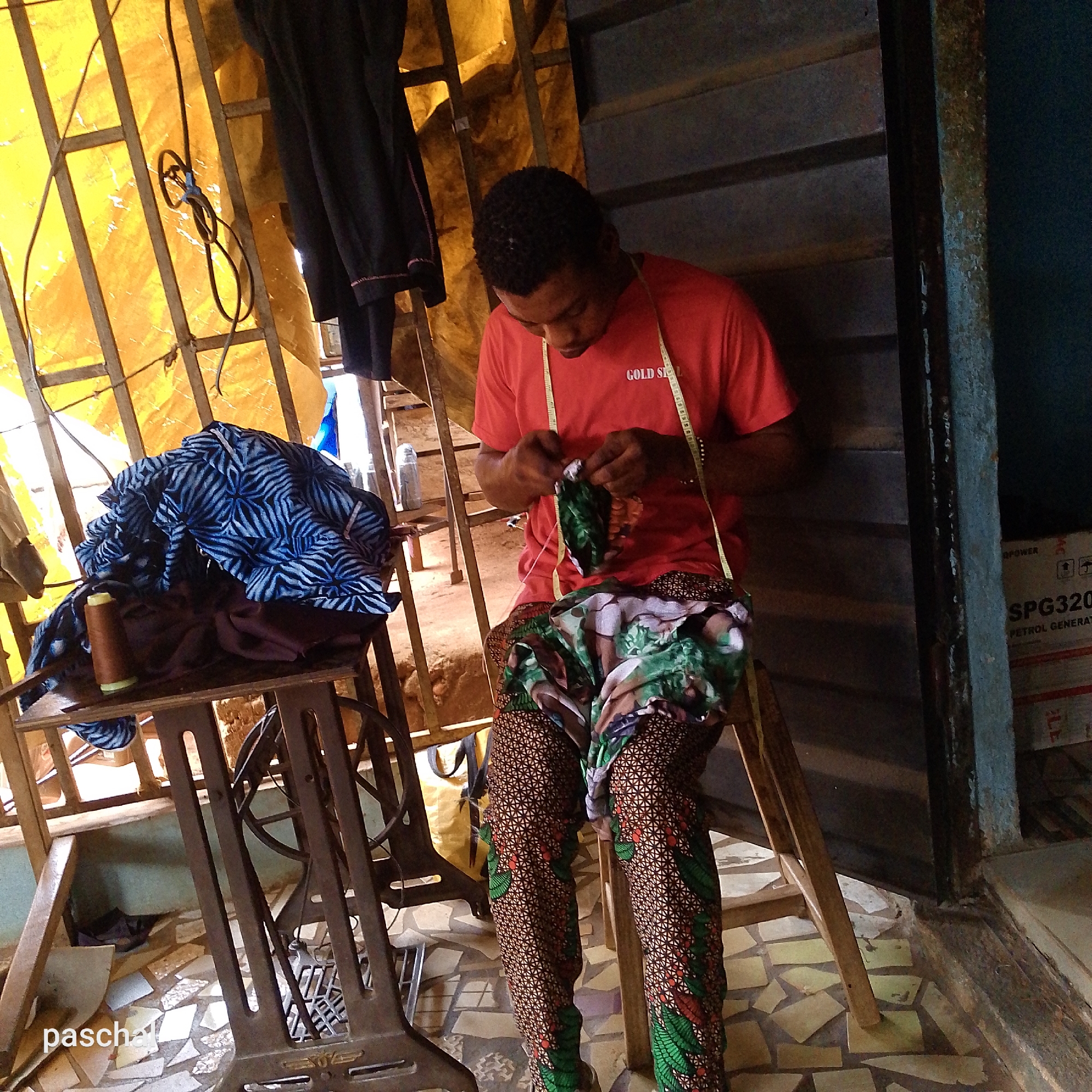 This full shot captures a young man diligently engaged in hand-sewing, likely in a tailoring workshop or stall in Ukwu-Abwa, Nigeria. He is seated on a simple wooden stool, wearing a red t-shirt with "GOLD S" visible on the chest, and vibrant trousers made of African print fabric featuring a geometric pattern with green and red foliage. A yellow measuring tape is draped around his neck.
The man is focused on a small piece of green and black patterned fabric that he is carefully stitching by hand. To his left, on a small wooden table, rests a pile of blue and white patterned fabric and a spool of brown thread. This table is integrated into the cast-iron base of what appears to be a vintage treadle sewing machine.
The setting features a structural framework of metal bars, partially covered by a translucent yellow tarp that diffuses the light, suggesting an open-air or semi-enclosed workspace. A dark, corrugated metal wall or door forms the background on the right side. A black garment is hanging from the metal frame in the upper left. The floor is an eclectic mix of irregularly shaped, light-colored tiles.
In the far right background, a white box is partially visible with the text "POWER", "SPG320", and "PETROL GENERATOR" printed on it, indicating a generator box. A tangled black electrical cord hangs from the overhead structure. The scene portrays a focused craftsman at work, immersed in a functional, community-level textile environment. The watermark "paschal" is visible in the bottom left corner.