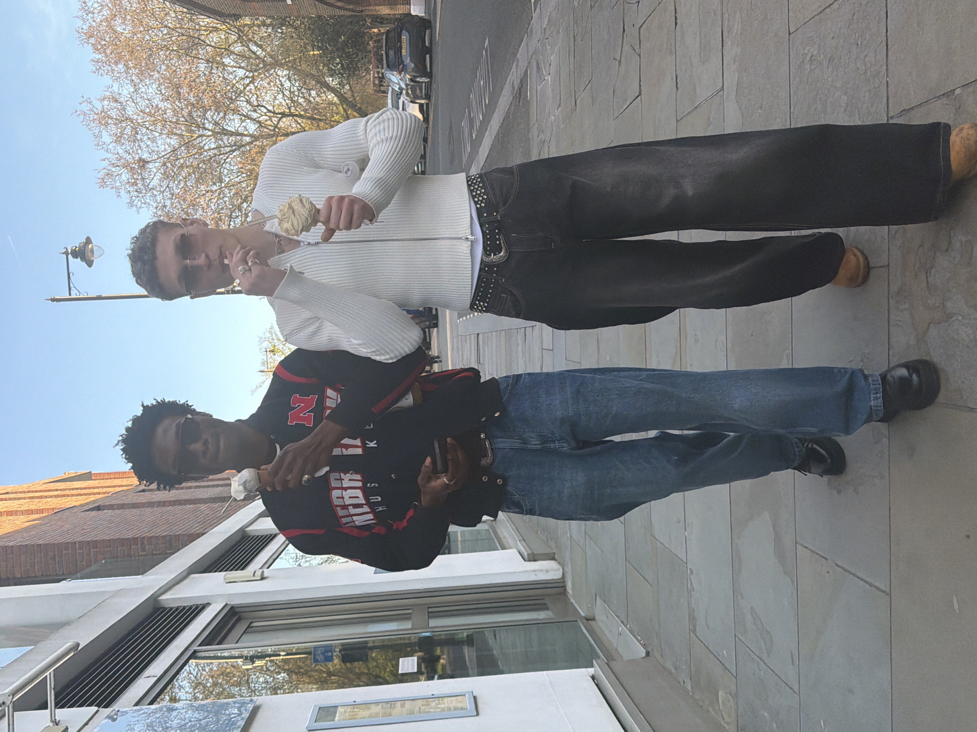 Two men stand on a paved sidewalk. The man on top wears a white zip-up sweater, black jeans with a studded belt, sunglasses, and light brown shoes; he is holding something white in his hand. The man below wears a black and red jacket with "NEBRASKA HUSKERS" on it, blue jeans, black shoes, and sunglasses; he also holds something white. The background includes a building with glass windows, a street with cars, bare trees, a street lamp, and a clear, blue sky. The location is London, United Kingdom, as indicated by the street markings and architectural style.