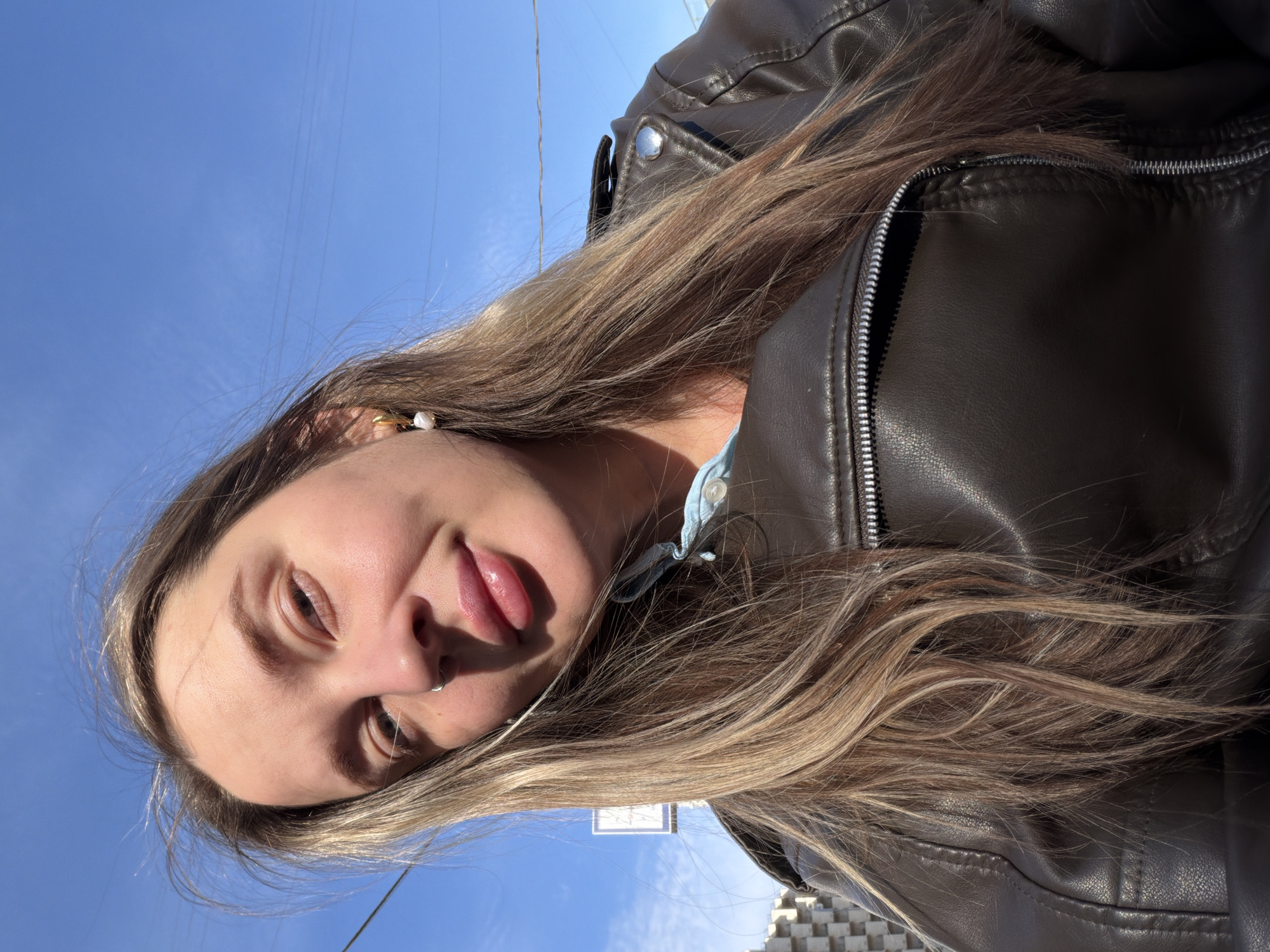 A young woman is pictured from a low angle with a blue sky in the background. She is wearing a dark brown leather jacket and has long, blonde hair with darker roots. She has fair skin and brown eyes. She has a nose ring in her left nostril and a pearl earring in her left ear. She is sticking her tongue out slightly. The background features a clear blue sky with some faint, white clouds. Although, I cannot confirm the city based on the provided image.