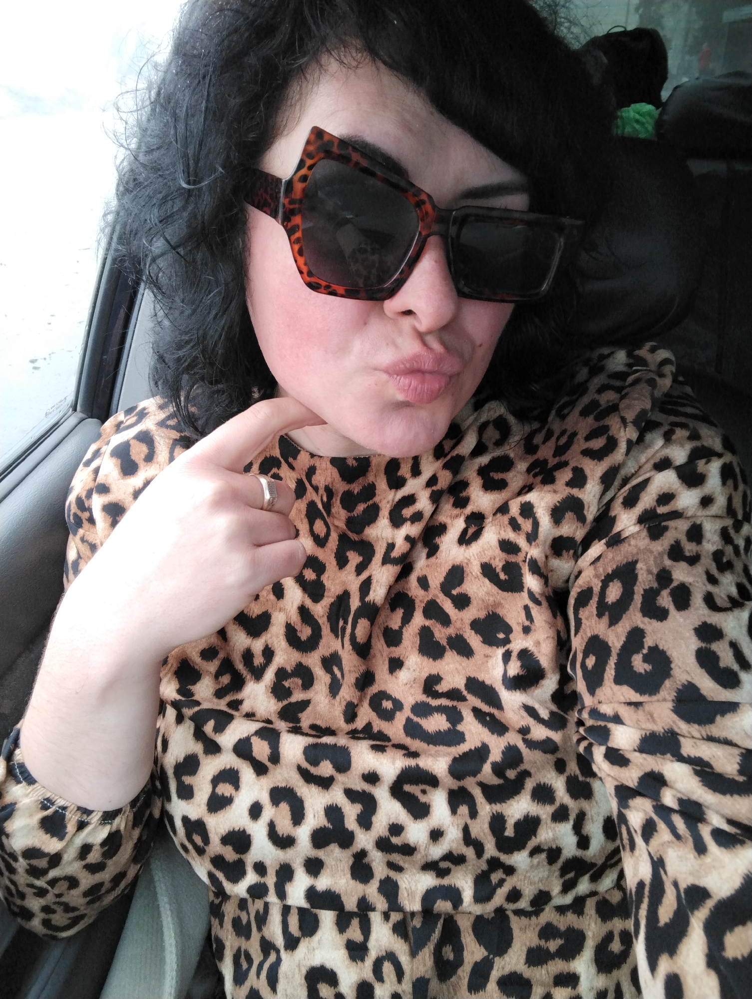 A woman with dark curly hair and bangs is posing for a selfie inside a car. She is wearing large, square sunglasses with a brown and black leopard print frame. She is also wearing a leopard print top. Her lips are pursed in a kiss. Her left hand is raised, with a finger touching her chin. She is wearing a silver ring on her left ring finger. The interior of the car is visible, with the door frame and seat visible. The background appears to be outside the car, but it is out of focus. There is no information available to determine if this image was taken in Votkinsk, Russia.