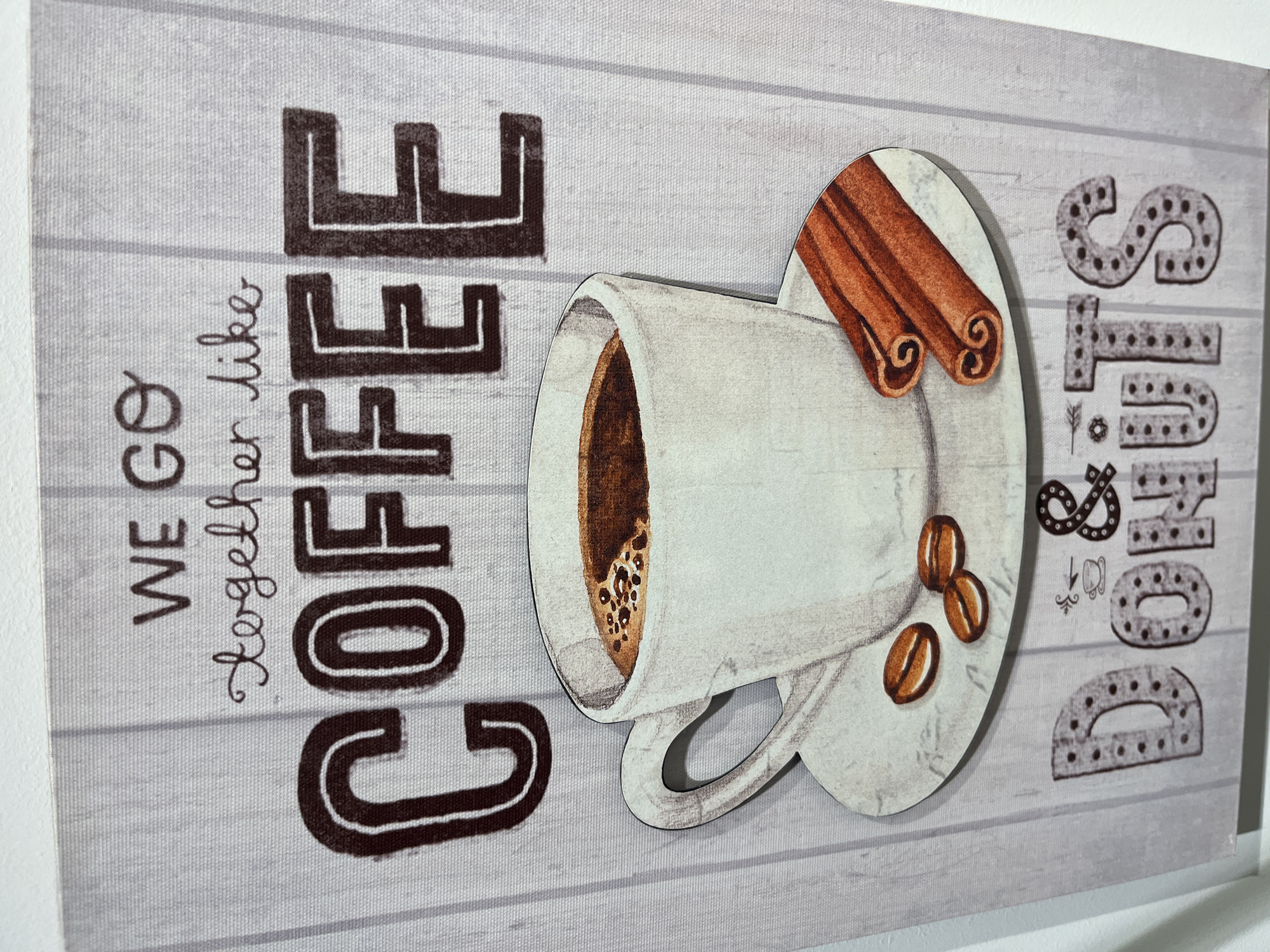 A decorative sign featuring the text "WE GO TOGETHER LIKE COFFEE & DONUTS" is displayed. The sign has a rustic, wood-plank background. The word "COFFEE" is written in large, bold, dark brown block letters, with "CO" on the left and "FFEE" on the right. Below "COFFEE" is a white mug filled with dark brown coffee, set on a saucer. Three coffee beans and two cinnamon sticks are placed on the saucer. The words "DONUTS" are written in smaller, dark brown block letters with a dotted pattern, separated by an ampersand symbol '&' and a small illustration of a coffee cup with steam. The sign is likely a decorative item for a cafe or home, and its specific location in Kharkiv, Ukraine, cannot be determined from the image itself.