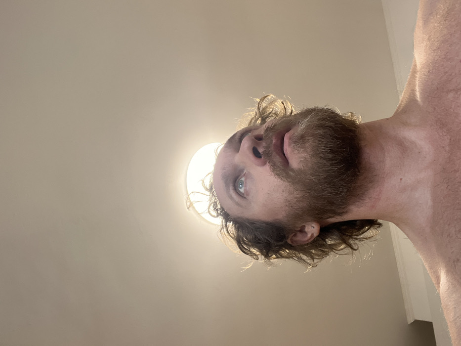 The image displays a close-up, low-angle perspective of an adult male indoors. The individual has light brown, curly hair that appears disheveled, and a full, reddish-brown beard. His light-colored eyes are directed upwards and slightly to the right, with his mouth slightly ajar. A small silver stud earring is visible in his left earlobe. He is shirtless, with his shoulders and upper chest visible. Directly above and slightly behind his head is a circular, bright white light fixture, providing illumination. The background consists of a plain, light-colored ceiling and a segment of a light-colored wall with white crown molding or trim on the right side. The scene is located in Saint Petersburg, Russia.