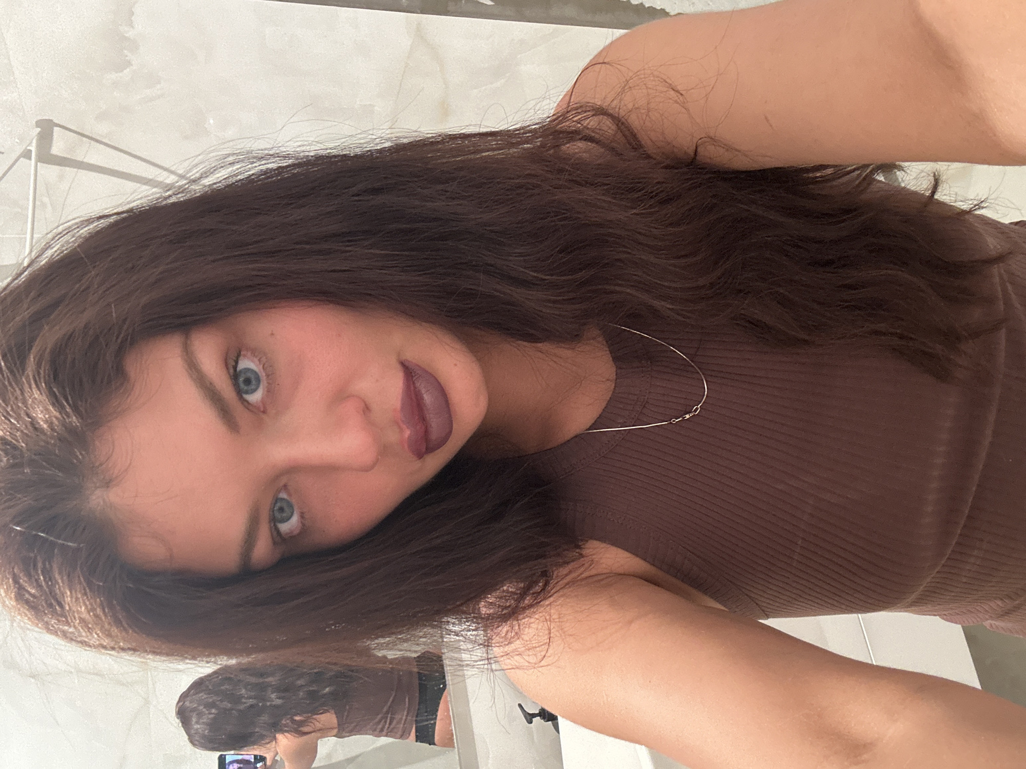 A woman with long brown hair, blue eyes, and wearing a brown top is taking a selfie in a bathroom. The background includes white tiles and a sink. A reflection of the woman taking the picture with her phone can be seen in a mirror. There is no evidence to confirm the location is in Khimki, Russia.