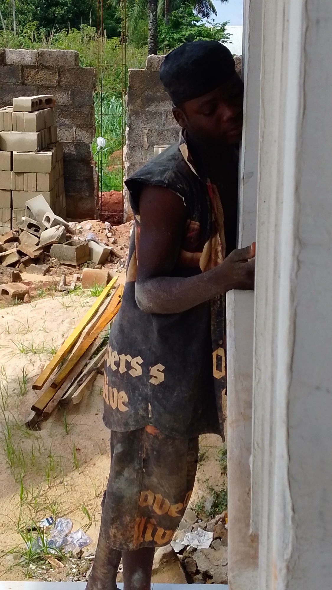 A man wearing a black cap, sleeveless shirt with the text "Diners Survive" and shorts, stands holding a white concrete frame. His clothes and skin are covered in dirt or dust. In the background, there is a partially constructed brick wall, a pile of bricks, and some wooden planks. The ground is sandy with scattered debris. The image was likely taken in Enugwu-Ukwu, Nigeria.