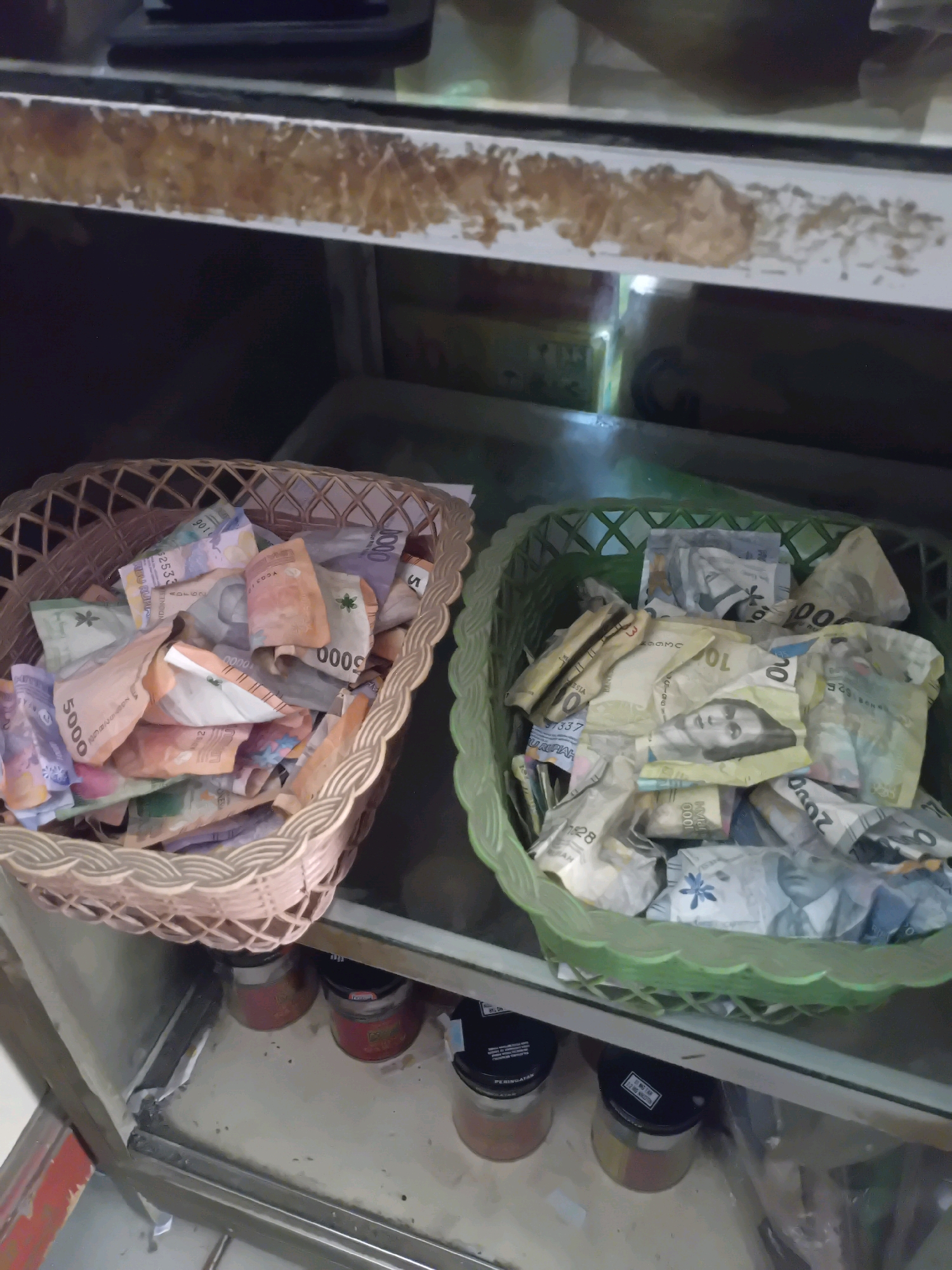 Two woven plastic baskets filled with Indonesian Rupiah banknotes are placed on a shelf. The basket on the left is light brown, and the one on the right is green. The banknotes appear to be of various denominations, including 5000, 1000, and potentially others. Some banknotes are folded or crumpled. The shelf is made of metal and glass, and appears to be part of a display or storage unit. Below the shelf, three small glass jars with red and yellow labels are visible. The overall setting suggests a commercial or retail environment. The presence of Indonesian currency indicates the location is likely in Indonesia, and potentially the city of Pekanbaru, Indonesia, if that information is contextual to the image. There are no people or animals visible in the image. The lighting is dim, with some artificial light illuminating the scene.
