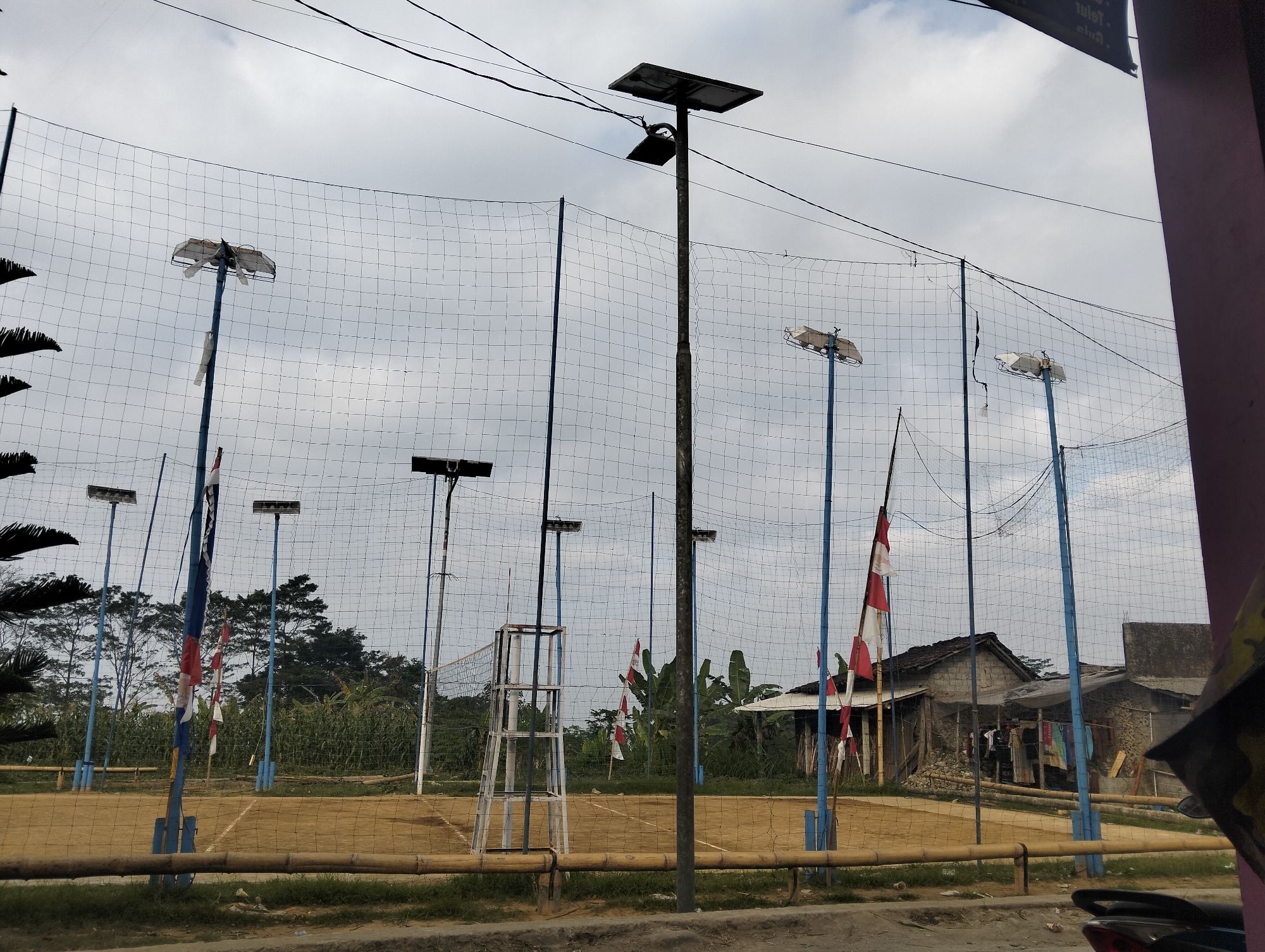 The image shows an outdoor volleyball court in Ropoh, Indonesia, under a cloudy sky. The court surface is made of tan-colored dirt with bamboo poles outlining the boundaries. A net is strung across the court, supported by blue poles. Several tall poles with lights and solar panels are positioned around the perimeter of the court, suggesting it can be used at night. Two Indonesian flags, red and white, are visible flying on poles. In the background, there are trees, dense vegetation including banana plants, and a few modest buildings with corrugated metal roofs and concrete walls. A white volleyball net stand is visible on the court. The scene appears to be a local recreational facility.
