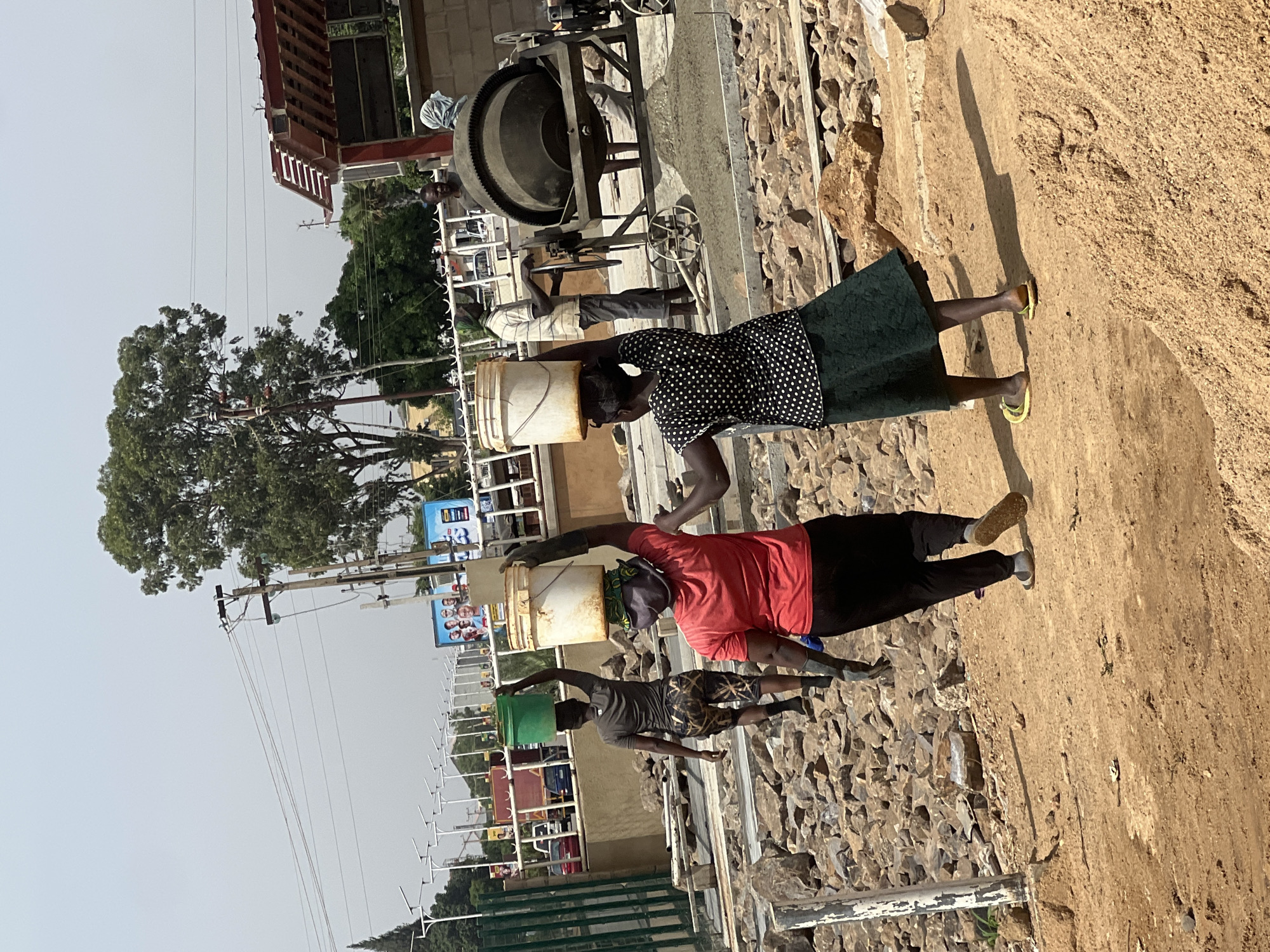 Three people are carrying buckets on their heads near a construction site in Barakin Lerawi, Nigeria. A woman in a black and white polka dot top and green skirt is closest to the foreground, followed by a person in a red shirt and black pants, and then a person in a gray shirt and patterned shorts. Each person carries a bucket, and the buckets appear to be of varying colors and sizes. They are walking on a dirt path beside a pile of rocks and stones. A cement mixer is located on a slightly elevated platform. Buildings, trees, streetlights, and utility poles are visible in the background. The sky is overcast.