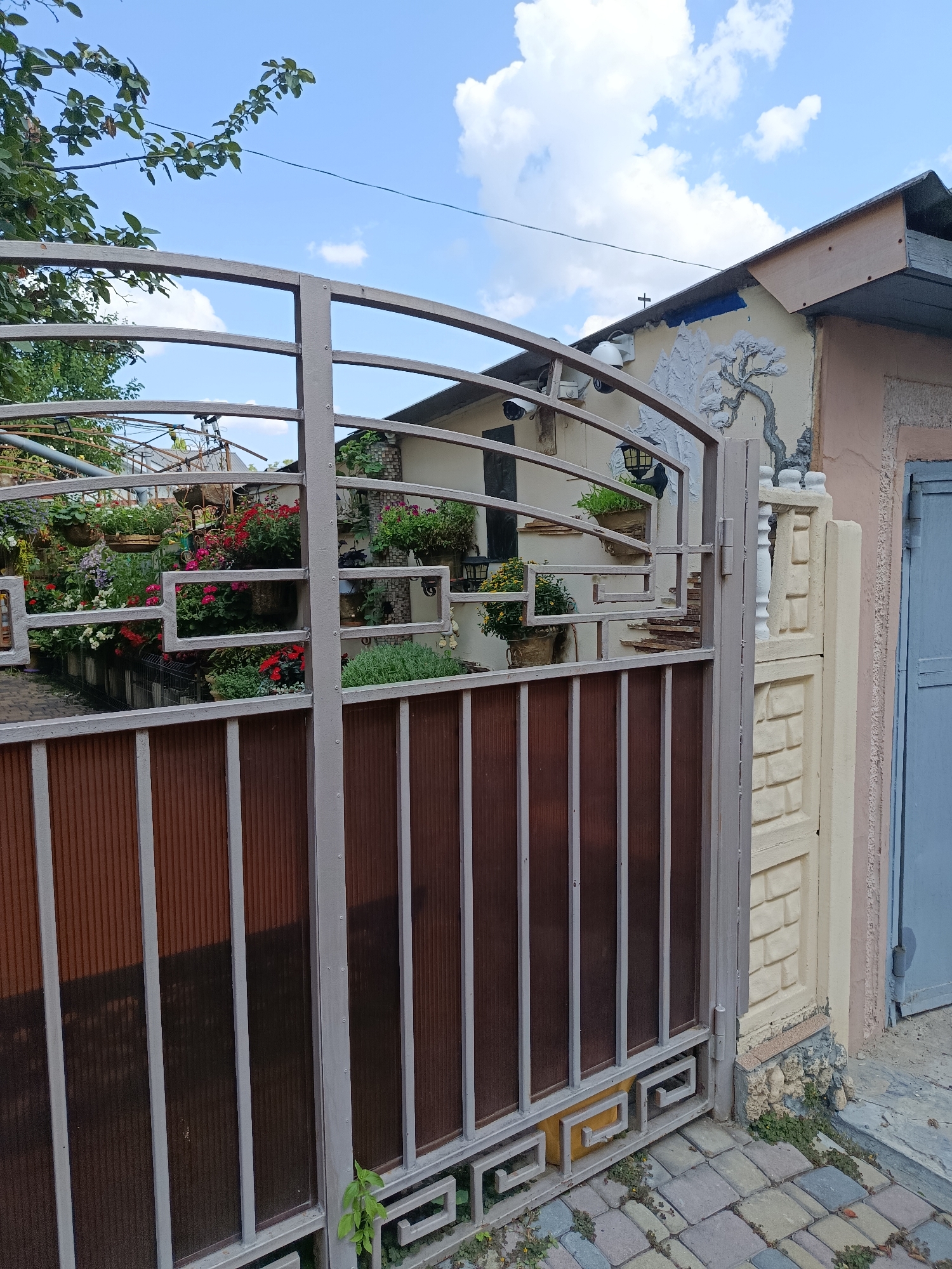 The image depicts a closed, ornate metal gate with brown tinted panels, situated in front of a building. Beyond the gate, a garden area is visible, filled with numerous potted plants, primarily flowering varieties in shades of red, pink, and white, along with greenery. The building behind the garden appears to have decorative elements on its exterior wall, including a relief of a tree and mountains. Security cameras are mounted on the wall. A cobblestone pathway is visible in the foreground leading up to the gate. To the right of the gate, a portion of a building with a blue door is visible. The sky is blue with scattered white clouds. The scene appears to be outdoors, in a residential or garden setting, likely in Chișinău, Moldova, given the context of the query. No people are present in the image.