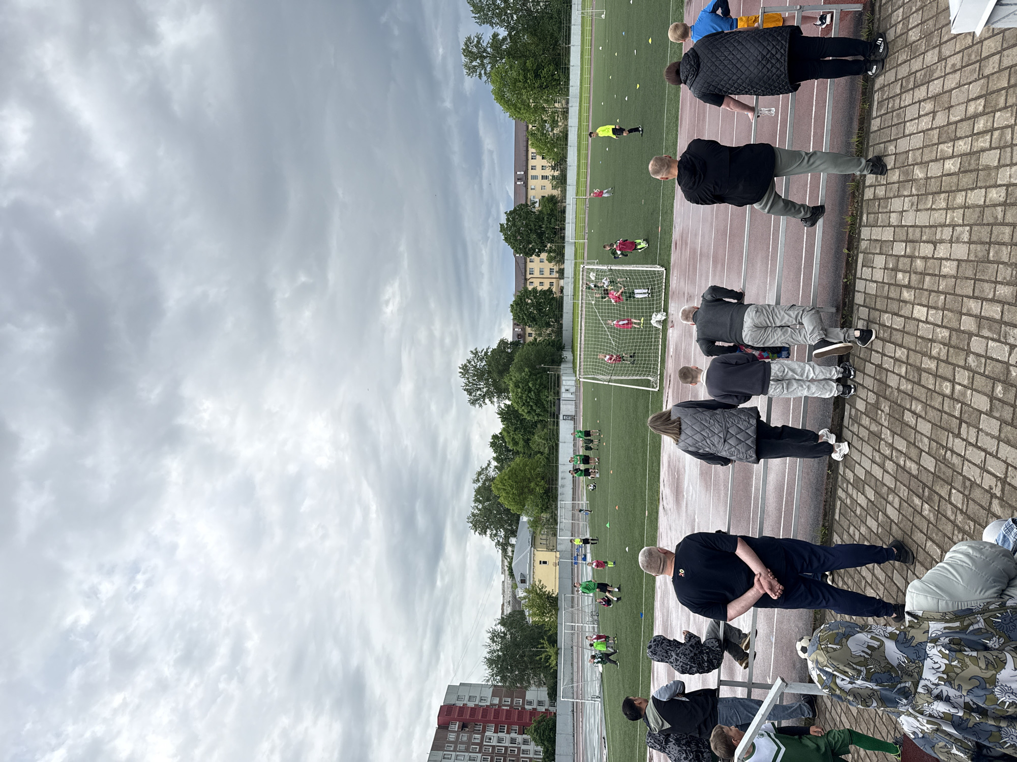 The image depicts a soccer training session taking place on a green artificial turf field. Players in green and red uniforms are engaged in drills around two soccer goals. An athletic track, marked with lanes, surrounds the field. A group of spectators, including adults and children, are standing along the track observing the training. A brick wall runs along the edge of the track. In the background, there are buildings and trees, including a red brick apartment building. The sky is overcast. The location is Veliky Novgorod, Russia.