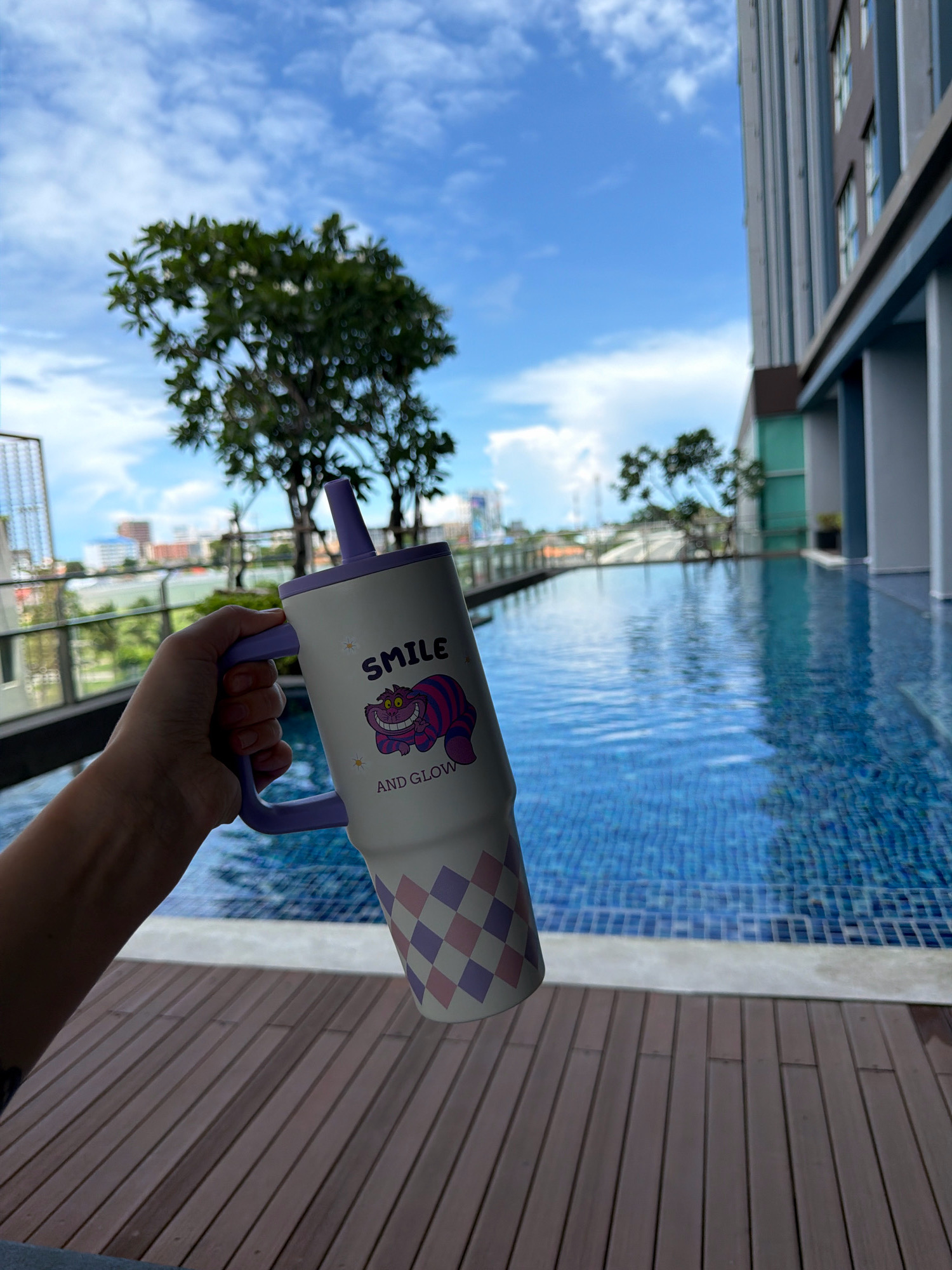 An overhead view shows a person holding a white insulated mug with a purple handle and straw. The mug is decorated with a purple and pink checkered pattern at the bottom and a grinning cat, labeled "SMILE AND GLOW," at the top. The mug is held in front of a swimming pool with blue tiled water. In the background, a tall building with grey vertical accents and windows stands to the right, while a lush green tree and other buildings are visible on the left. The scene is set against a bright blue sky with scattered white clouds, suggesting it is daytime in Hua Hin City Municipality, Thailand.