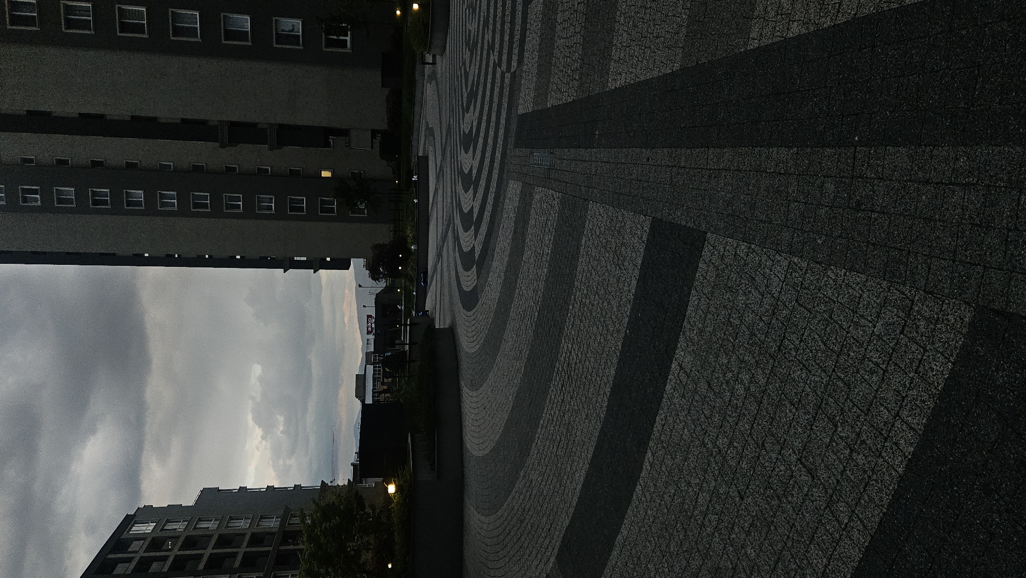 The image shows the central square in Plovdiv, Bulgaria, featuring a distinctive pavement design of concentric circles and radiating lines made of alternating light and dark grey paving stones. Adjacent to the square is a tall, multi-story building with rows of windows, captured against a backdrop of a cloudy sky. The scene has a cool, subdued lighting, possibly indicating dusk or an overcast day. In the background, beyond the immediate square and building, are hints of other buildings and possibly a sign, suggesting a wider urban environment. No people are visible in the frame.
