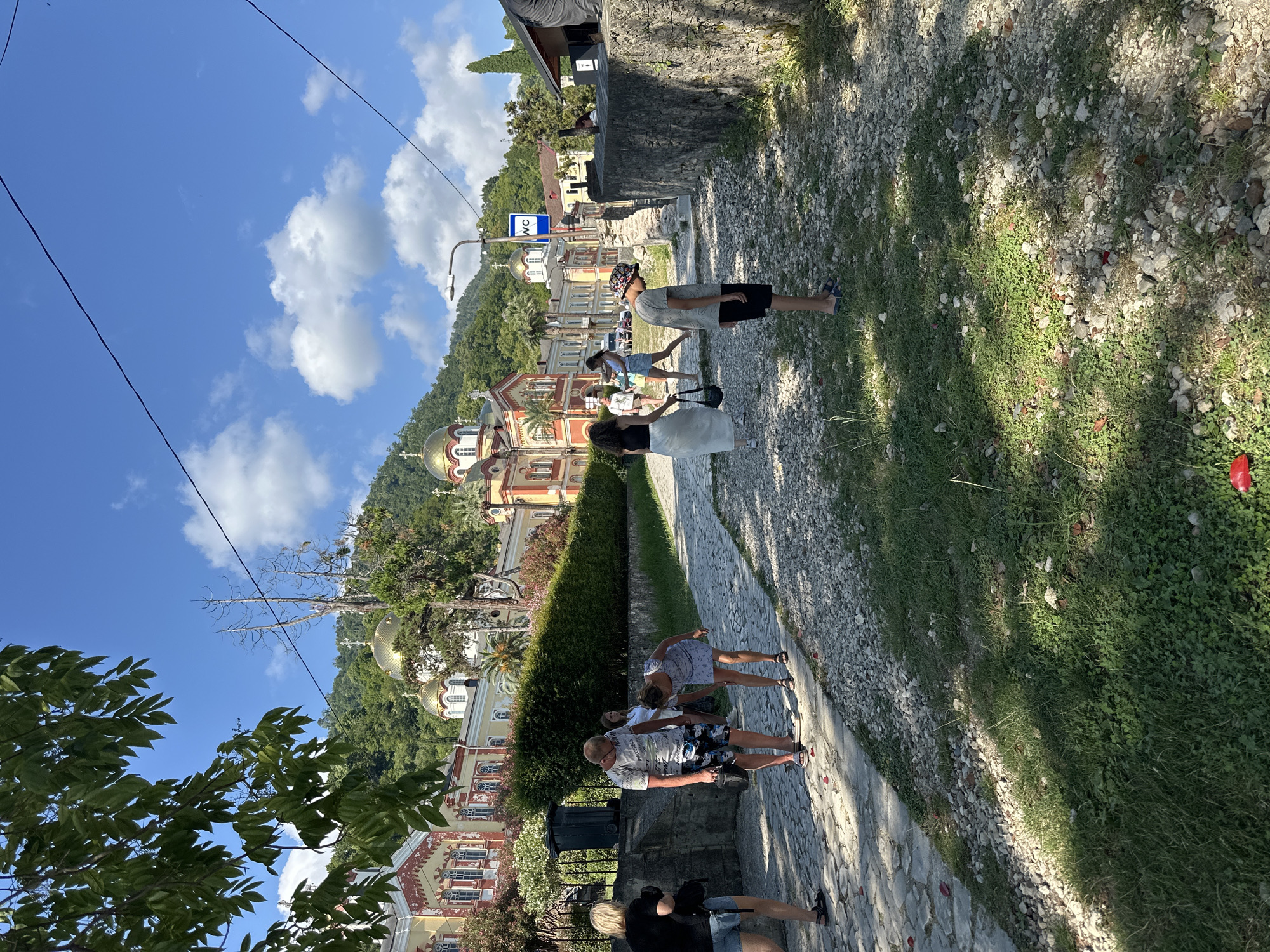 The image shows a sunny day at the New Athos Monastery in Abkhazia. Several people are walking on a cobbled path. The monastery, with its distinctive golden domes and colorful architecture, is visible in the background, nestled against a lush, green hillside. A WC sign is mounted on a lamppost. The path is lined with a low stone wall on one side and a dense hedge on the other, with trees and foliage bordering the scene. The ground is a mix of cobblestones and gravel, with patches of grass.
