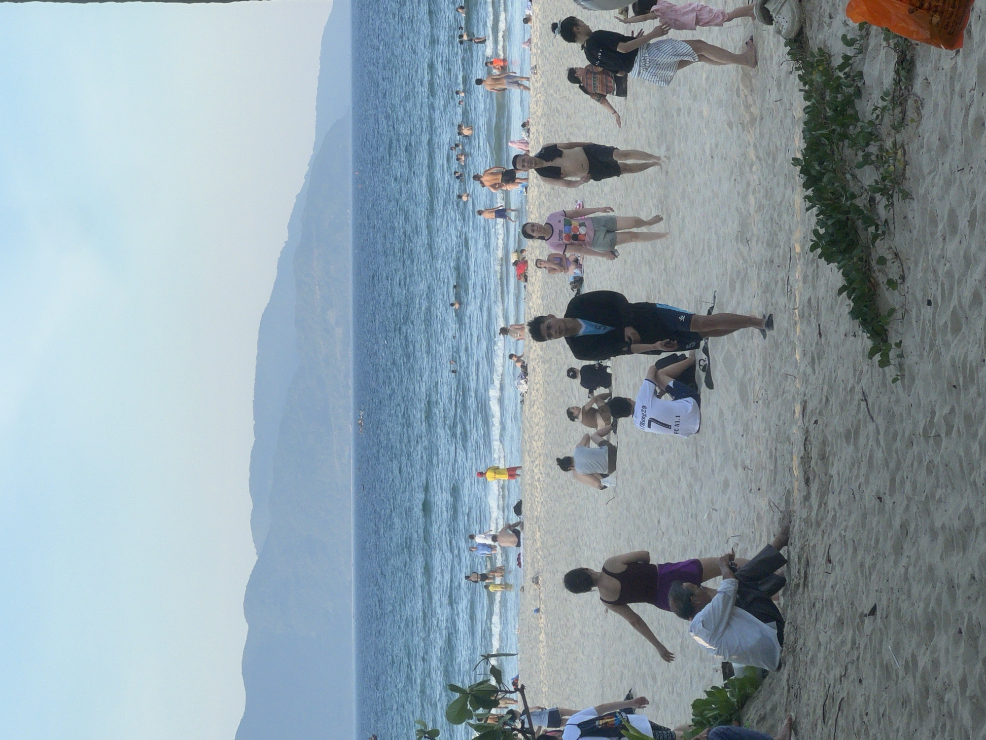 This media file captures a lively scene at a beach in Đà Nẵng, Vietnam. The image is oriented vertically, with the ocean on the left and the sandy beach on the right.
**Subjects:** The beach is populated with numerous people, most of whom are dressed in beachwear, indicating a casual atmosphere. There are men, women, and possibly children enjoying the seaside. Some are wading in the shallow waters, while others are walking along the shore or relaxing on the sand. In the foreground, a few individuals are more prominently featured, engaged in various activities.
**Activity/Event:** The primary activity is a day at the beach, with people swimming, sunbathing, walking, and socializing. The presence of many people suggests a popular beach on a pleasant day.
**Notable Details:** The time of day appears to be late afternoon or early evening, given the soft light and the angle of the shadows. The weather seems to be good, with clear skies and calm waters. The background shows a hazy view of mountains, a common characteristic of the Đà Nẵng coastline. The sand is light-colored and textured, with some vegetation growing towards the right side of the frame. Emotions conveyed are generally relaxed and joyful, typical of a beach outing.
**Visible Text:** Some text is visible on a white t-shirt worn by a person in the middle right of the image. It appears to read "Mừng 29" and "FC A.I.I". There is also some faint text on an orange object in the upper right corner, but it is illegible.