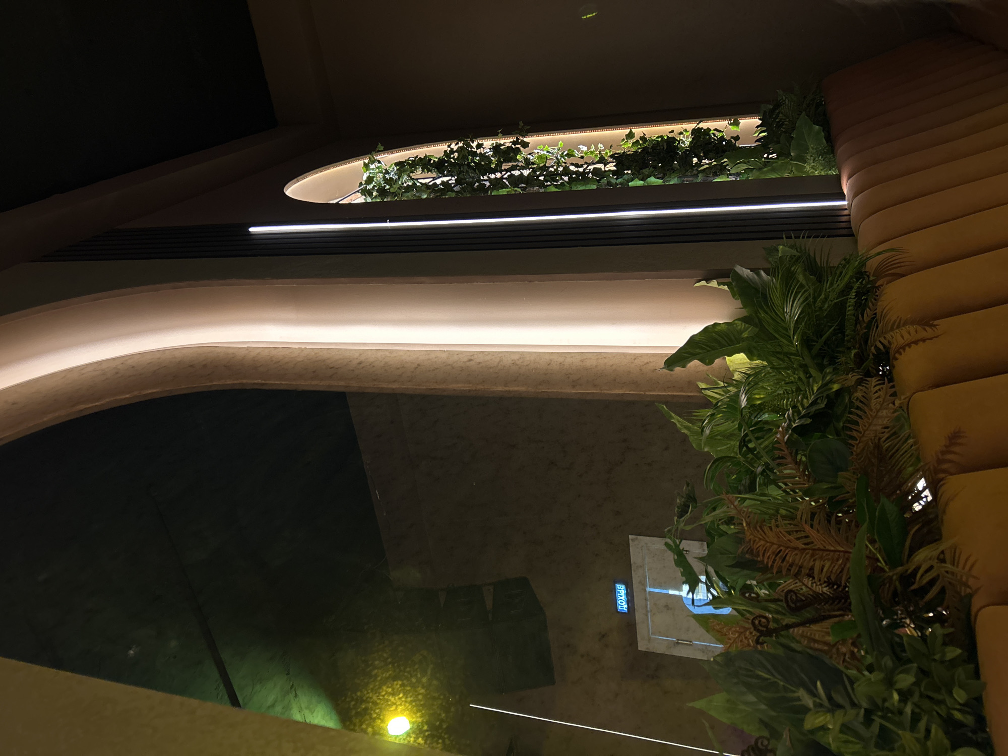 The image displays a dimly lit, modern interior space, likely a restaurant or lounge, characterized by warm ambient lighting and integrated greenery. The scene suggests evening or nighttime.
Key elements include curved architectural forms and prominent linear light strips that illuminate the ceiling and wall contours. Along the upper wall, a narrow, recessed planter filled with lush green foliage, possibly ivy, is illuminated from within, casting a soft glow on the plants. To the right, a textured wall or padded seating area with horizontal ridging, in a tan or ochre hue, adds a soft, organic feel.
The lower left quadrant features a large, dark, reflective surface, which could be a polished floor or a decorative body of water, mirroring the overhead architecture and a distinct bright yellow light source. A variety of tropical and fern-like plants are strategically placed throughout the space, enhancing the natural aesthetic. A small digital display is visible on a light-colored panel, showing the Russian word "ВЫХОД" (EXIT) in glowing blue text, and possibly a time, like "20:01". No people are present, and the overall atmosphere appears calm and inviting, designed for relaxation or dining.