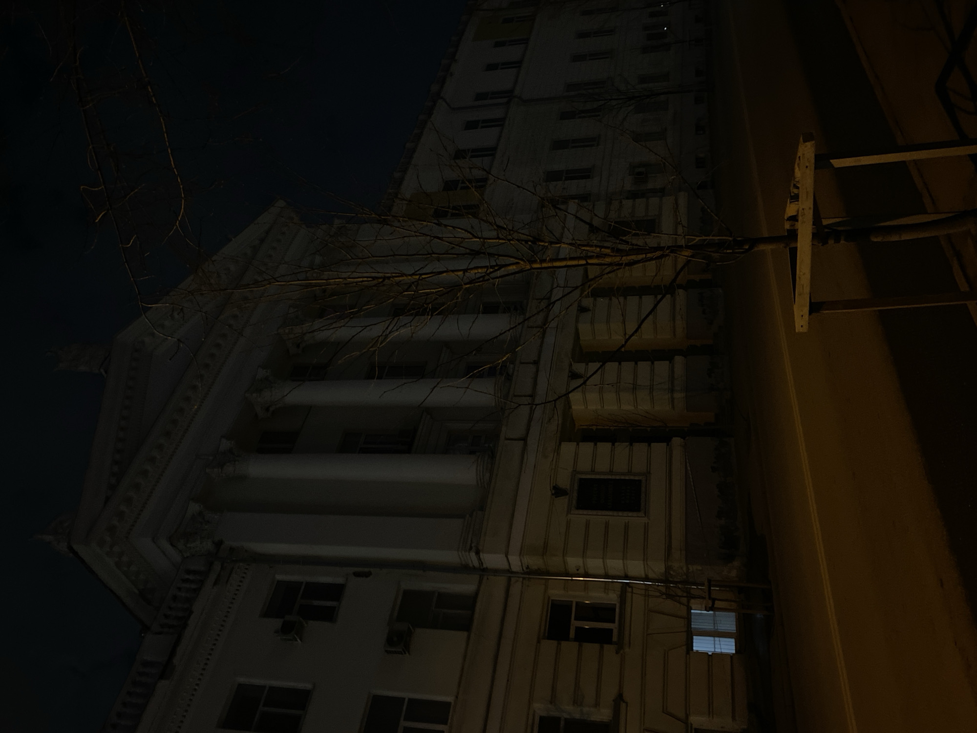 A low-angle, nighttime photograph depicts a tall, multi-story building in Kazan, Russia. The building's architecture features classical elements such as columns and decorative cornices. Many windows are visible, some with visible air conditioning units. A bare tree branch stretches across the building's facade. To the right of the building, a dark, vertical wall is partially visible, with a metal structure attached. The image is generally dark.