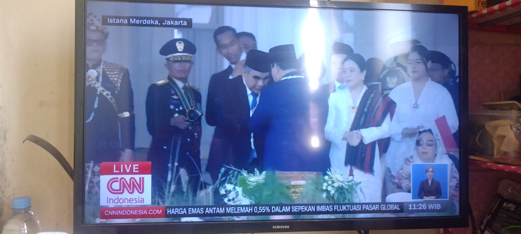 The image shows a television screen displaying a news broadcast from CNN Indonesia. The broadcast features a ceremony at the Istana Merdeka in Jakarta, Indonesia. Several people are visible in the broadcast, including men in military uniforms and formal attire, and women in traditional Indonesian clothing. The news ticker at the bottom of the screen displays "CNN Indonesia" and a headline in Indonesian that translates to "Gold Prices Weakened 0.55% Due to Global Market Fluctuations" along with the time "11:26 WIB". The television is placed in what appears to be a domestic setting, with a wall and part of a shelf visible in the background. A water bottle is also visible in the foreground on the left side of the image.