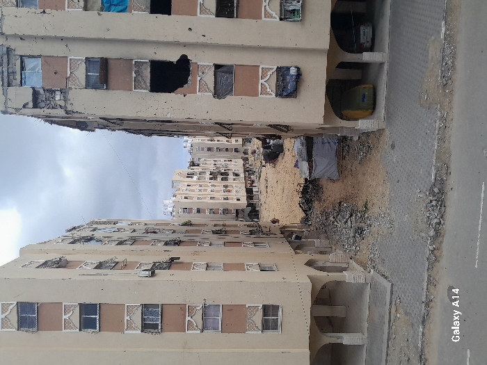 The image depicts a devastated urban scene in Khan Yunis, Palestinian Territory, characterized by extensive structural damage to multi-story residential buildings and a debris-strewn central clearing under an overcast sky.
In the foreground, a gray paved surface, possibly a road or plaza, is scattered with rubble and dirt. To the left and right, two multi-story residential buildings, predominantly light tan with brown accents, frame a central area. Both buildings feature decorative arched and patterned elements around their windows and arched ground-floor supports. The building on the left exhibits moderate damage, with several broken windows and minor pockmarks on its facade. The building on the right shows severe damage, including large, irregularly shaped holes penetrating several floors, exposed rebar, and numerous shattered windows, some covered with blue tarps or dark materials. Beneath the right building's arches, two vehicles are parked: one appears to be a light-colored sedan, and the other is partially obscured and covered.
Between these two primary buildings lies a wide, open, sandy clearing heavily covered with concrete rubble, twisted metal, and general construction debris. In the mid-ground of this clearing, a makeshift structure constructed from a white tarp is visible. Several small figures, appearing distant, are present in the clearing; one figure wearing a red top is discernible walking towards the left. In the background, beyond the clearing, more multi-story buildings, also appearing damaged, are visible.