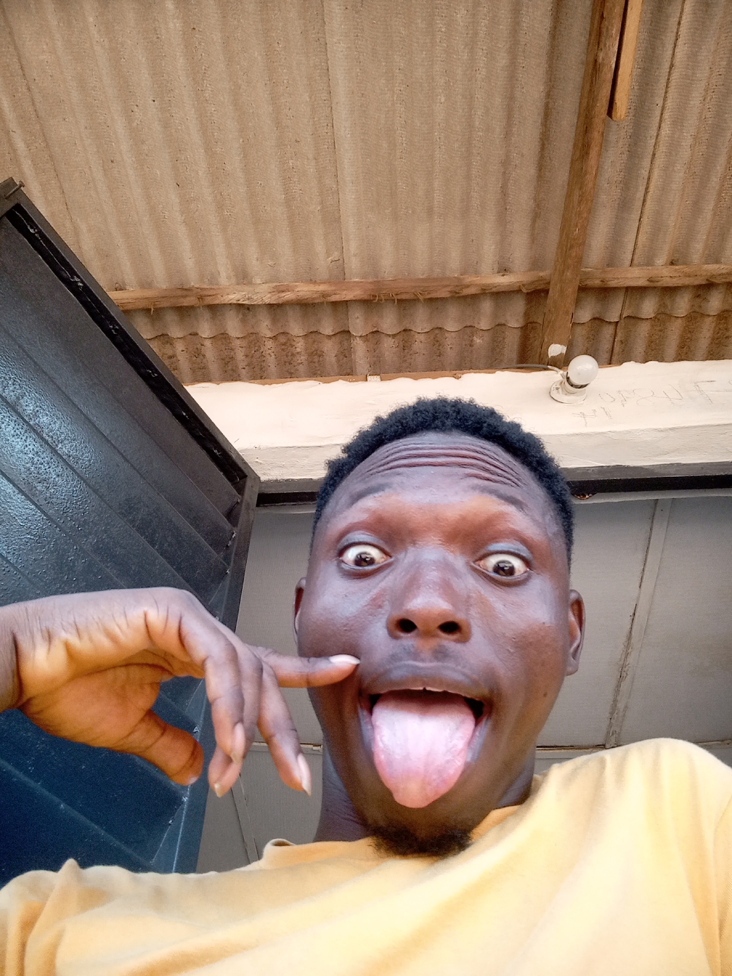 A young man with dark skin and short dark hair is featured in a close-up, low-angle selfie, making a playful expression. His eyes are wide open, and he is sticking out his tongue. His left hand is raised, with his index finger touching his cheek. He is wearing a yellow t-shirt.
The background reveals an indoor or sheltered outdoor setting. Above the man's head, a corrugated ceiling or roof structure made of a light brown or grey material is visible, supported by rough wooden beams. To the left, part of a dark, possibly black, louvered door or shutter is seen. To the right and behind the man's head, there is a white wall or beam with a simple, bare light bulb fixture attached. Faint, unreadable markings or text appear on this white surface near the light fixture. The lighting is bright and even, suggesting it is daytime. The overall mood of the image is lighthearted and humorous.