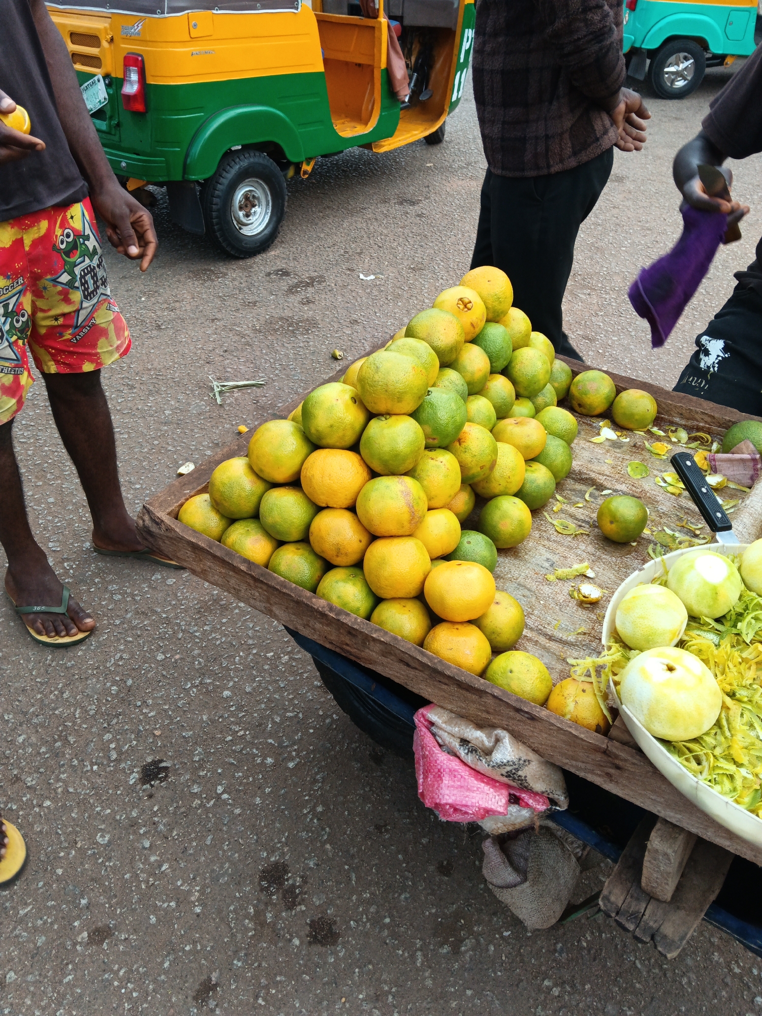 A street vendor in Bukuru, Nigeria, arranges a cart overflowing with oranges. The cart, made of wood, is positioned on a paved surface. The oranges, a mix of green and yellow, are piled high, with some already peeled and displayed in a white bowl, accompanied by shredded peel. A black knife rests on the cart, suggesting the preparation of the fruit for sale. In the background, a colorful three-wheeled auto-rickshaw, yellow and green, is visible, indicating a busy market or roadside setting. Two individuals are partially visible in the frame; one is wearing a dark shirt and patterned shorts, and the other is in a brown, checkered jacket. The scene is lit by natural daylight.