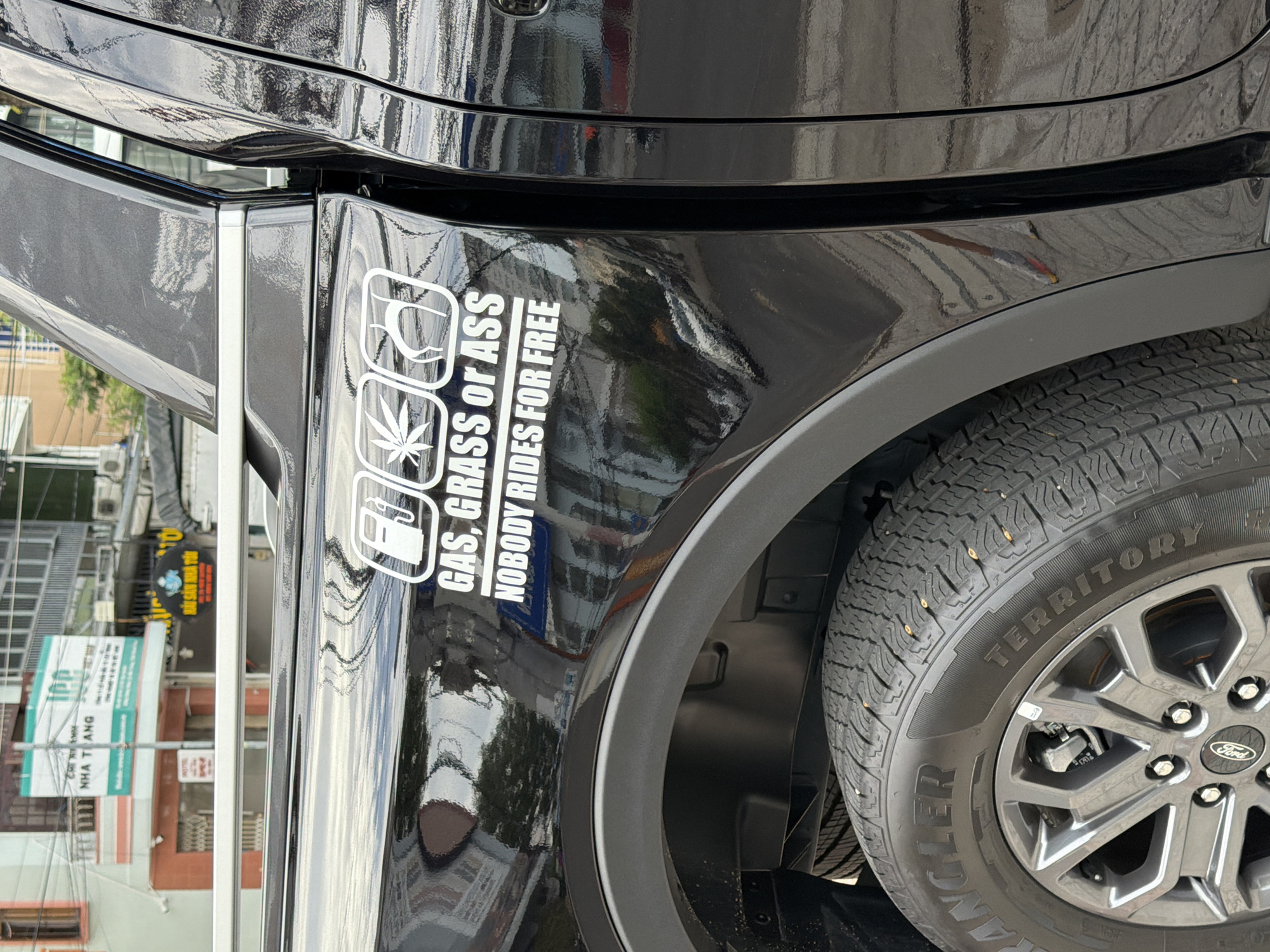 The image captures a close-up view of the rear passenger side of a black pickup truck parked on a street in Nha Trang, Vietnam. A white sticker with three icons and text is prominently displayed on the truck's body. The icons, from left to right, appear to be a gas pump, a marijuana leaf, and a pair of buttocks. The text below reads "GAS, GRASS or ASS" and "NOBODY RIDES FOR FREE". The truck's glossy black paint reflects the surrounding environment, including buildings and trees. In the background, signs for "IPC" and "NHA TRANG" are visible on a building, indicating the location. The truck's tire, a "WRANGLER TERRITORY" model, and part of its alloy wheel with a "Ford" emblem are also in frame. The street appears to be paved, and overhead wires are present. The overall scene suggests a typical urban street setting in Vietnam.