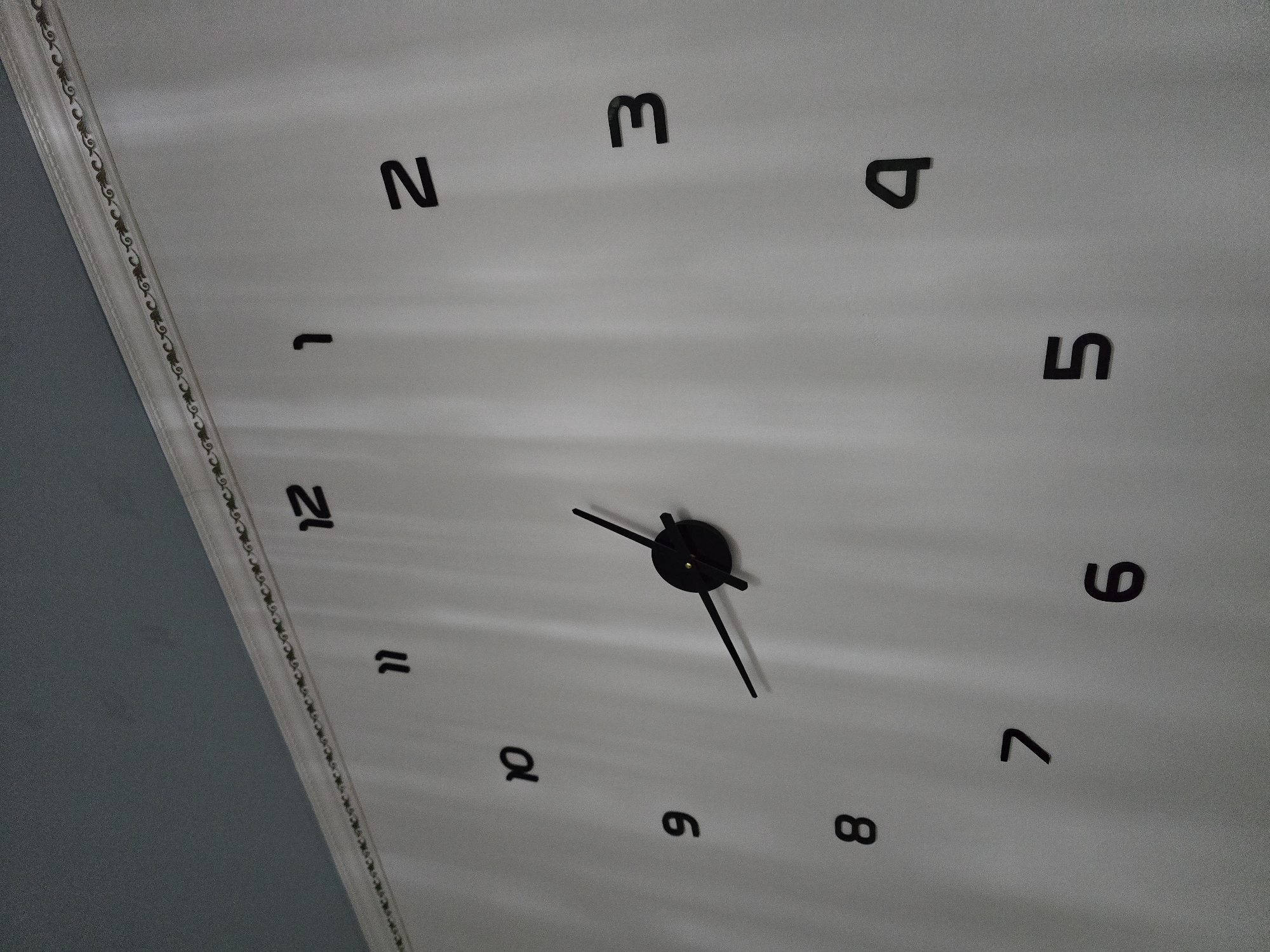 A black analog clock with oversized numbers is mounted on a white wall. The clock has black hour and minute hands, with the minute hand pointing to the "5" and the hour hand pointing slightly past the "10". The numbers on the clock are arranged in a circular pattern, with "12" at the top, "3" on the right, "6" at the bottom, and "9" on the left. The wall has a decorative border running along the top edge, and a grey ceiling is visible above the border. The scene is indoors and well-lit. The image does not depict any people, actions, or interactions. There is no information in the image to suggest its location in Qaratau, Kazakhstan.