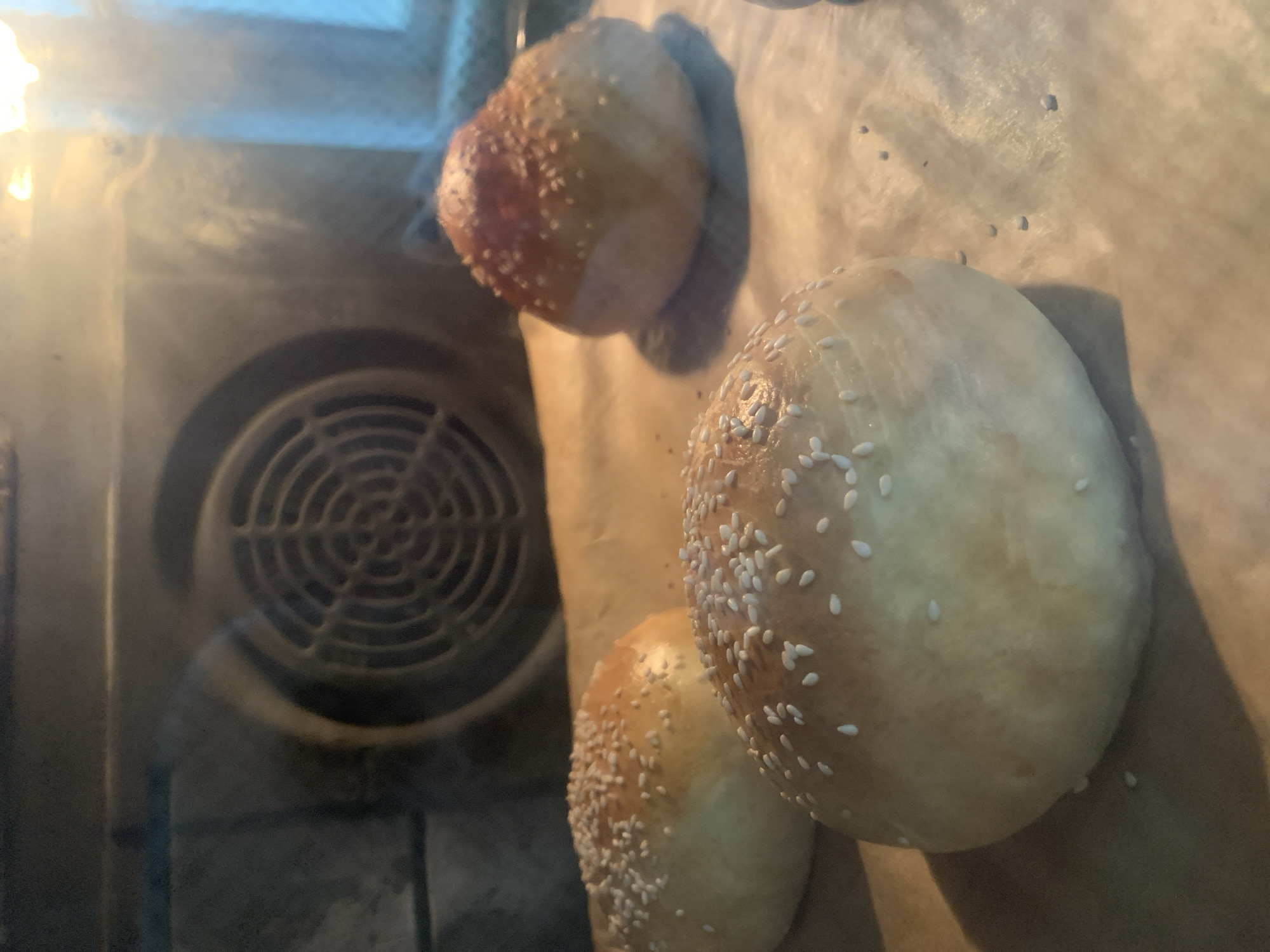 Four hamburger buns are baking on a parchment-lined baking sheet inside an oven. The buns are golden brown and sprinkled with sesame seeds. The oven fan is visible in the background. The scene is indoors.