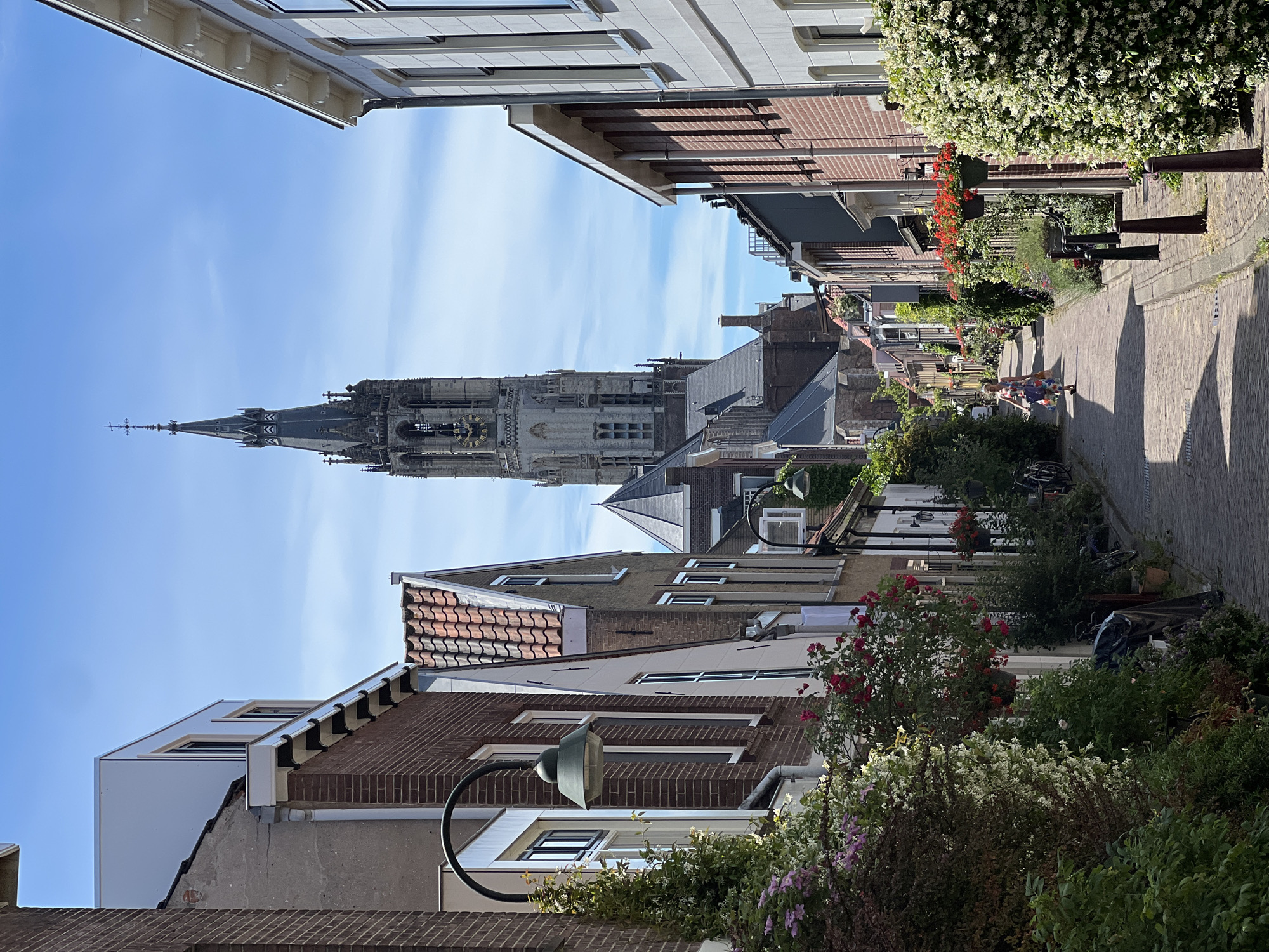 The image shows a narrow, cobblestone street in Delft, Netherlands, leading towards the towering spire of the Nieuwe Kerk. The street is lined with brick buildings of varying heights, many adorned with flower boxes and greenery, creating a picturesque and charming urban scene. A lone woman, wearing a colorful, flowing dress, walks away from the camera down the street. Bicycles are parked along the sides of the road, and street lamps are visible on some of the buildings. The sky is bright blue with scattered clouds, indicating a pleasant day. The perspective draws the viewer's eye towards the prominent church spire in the distance, a landmark of Delft.