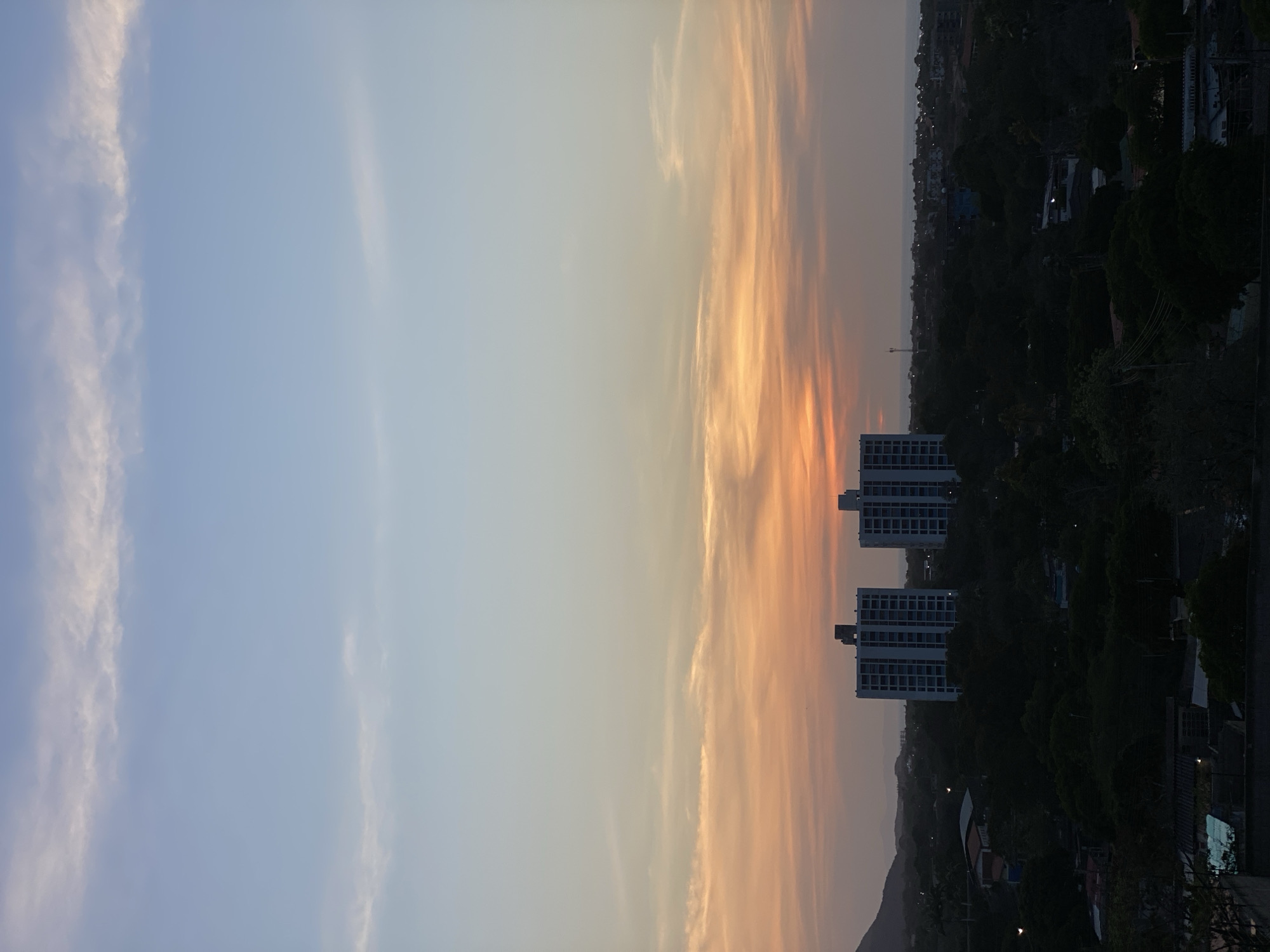 The image presents an evening view from Juan Griego, Venezuela. The sky transitions from light blue to orange-tinged clouds as sunset approaches. Two high-rise buildings stand amid dense trees and other buildings. Further in the distance, the silhouettes of hills frame the horizon.