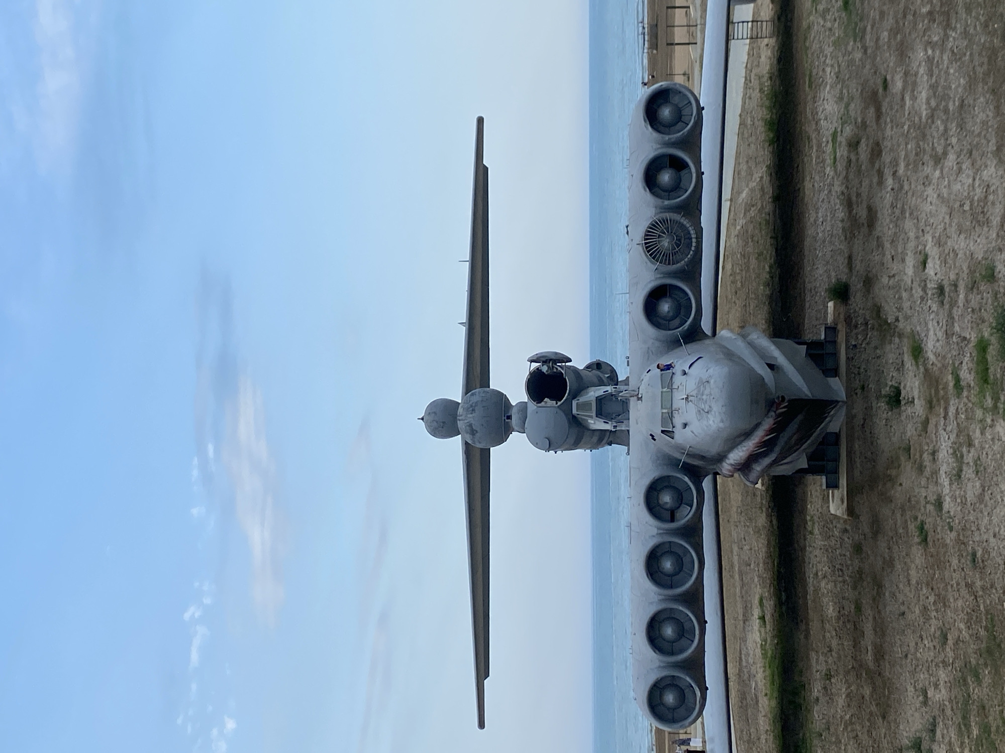 A front-facing, eye-level, full shot captures an imposing, gray, Soviet-era ekranoplan (ground-effect vehicle) resting on a sandy, dry dirt surface. The colossal aircraft features a distinctive design with a long, wide body and two expansive wings that stretch horizontally. Along the underside of the starboard wing, six circular engine nacelles are visible, with a seventh engine, a different design, positioned further outboard. The port wing mirrors this arrangement, though fewer engines are fully visible due to the angle. The nose of the ekranoplan is pointed and features a painted shark mouth graphic with sharp teeth. A tall, multi-tiered structure rises from the center of the aircraft's body, housing additional engines and what appears to be a cockpit area. The background is dominated by a vast, calm ocean stretching to the horizon, with a hazy, pale blue sky above, marked by thin, wispy clouds. The environment suggests a coastal location, possibly a museum or preserved exhibit.