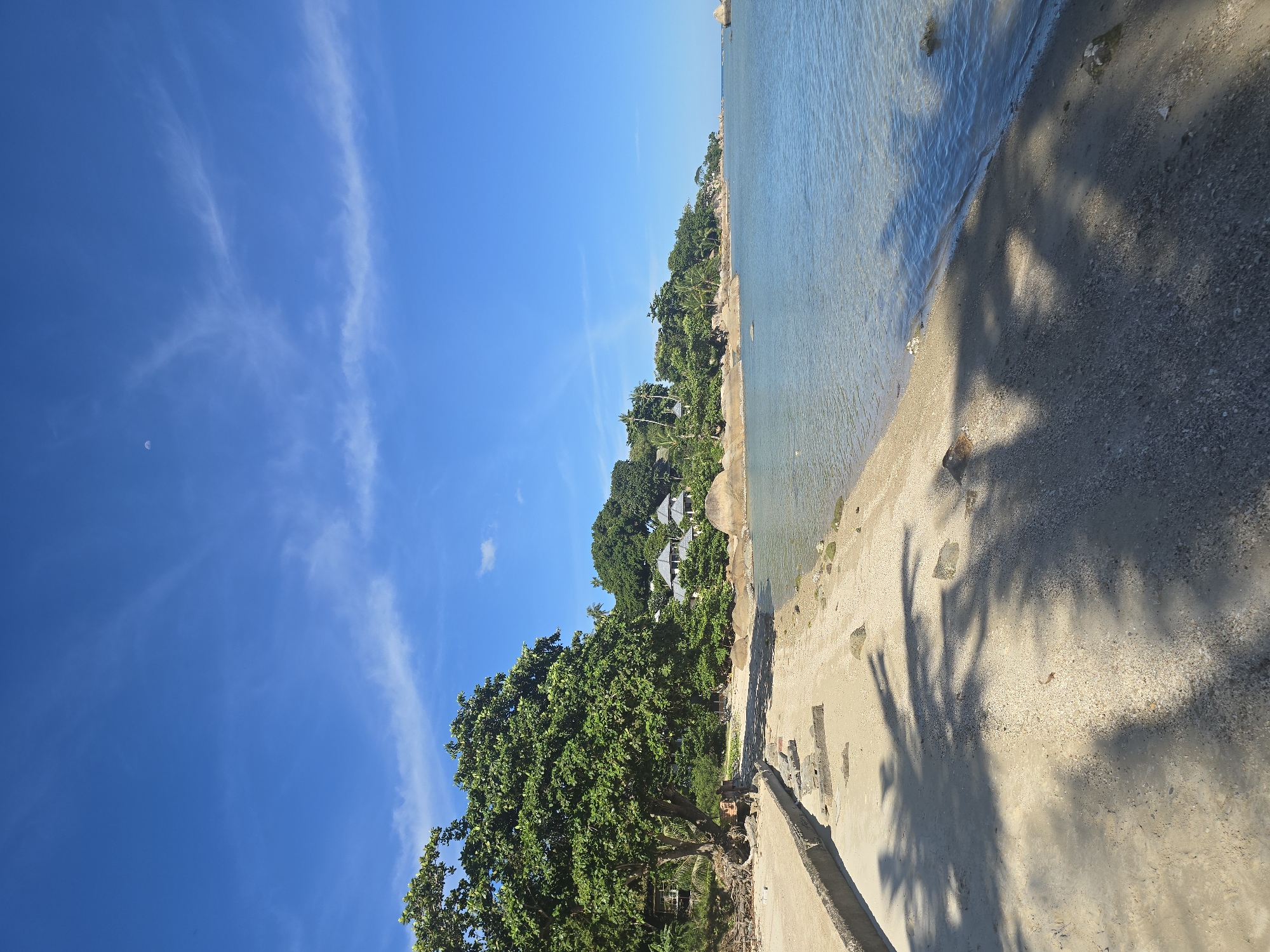 The image depicts a serene coastal scene with a clear blue sky and calm, light-colored water. A sandy beach lines the foreground, with shadows of palm leaves cast across it. To the left, a weathered concrete structure runs along the edge of the beach. Beyond the beach, lush green trees and several houses with grey roofs are visible, nestled against a backdrop of rocky outcrops and more vegetation. The water is shallow and transparent near the shore, gradually deepening to a richer blue further out. The horizon is clear, with a faint crescent moon visible in the upper portion of the sky. The scene conveys a peaceful tropical atmosphere.