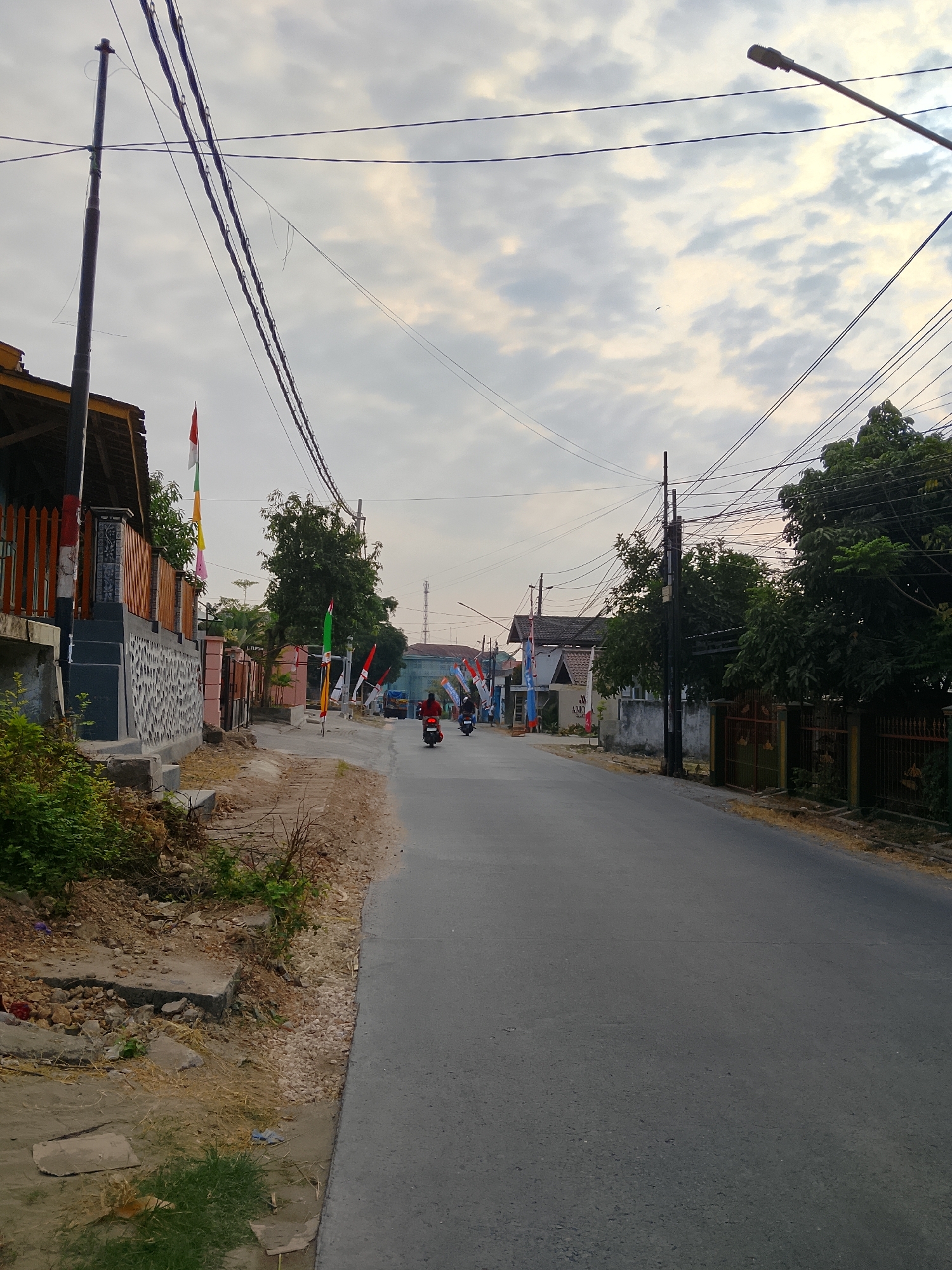 The image depicts a street scene in Ngelo, Indonesia, under an overcast sky. The road, paved with asphalt, is the dominant feature, curving slightly to the right. On the left side of the road, there is a sidewalk area with uneven terrain, featuring patches of dry grass, scattered rocks, and debris. Adjacent to the sidewalk is a residential property with a fence and a building with a wooden fence and a dark-colored wall. Various flags, predominantly red and white, are seen mounted on poles along the street and on the property.
On the right side of the road, there are more residential properties with fences, trees, and utility poles. A street light is visible at the top right of the image. Several electrical wires crisscross overhead.
In the middle of the road, two individuals on motorcycles are traveling away from the viewer. One motorcycle has a rider wearing a red shirt, and the other has a rider wearing a dark top. Further down the road, more flags are visible, suggesting a festive or commemorative occasion. In the background, other buildings and trees can be seen, and a faint silhouette of a tower or antenna is present on the horizon. The overall impression is a typical street in a residential area.